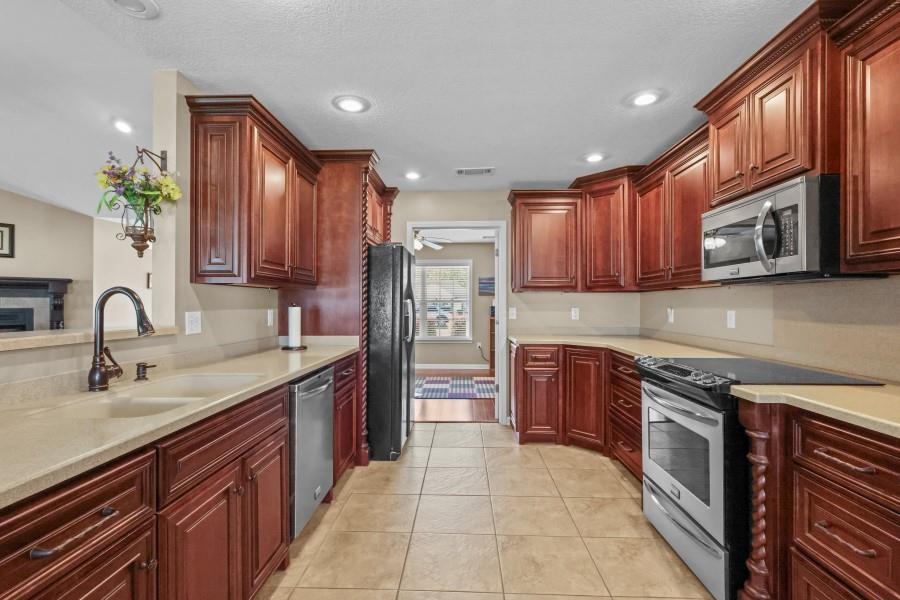Kitchen with reddish wood cabinetry, light stone counters, stainless-steel appliances, and a tiled floor.