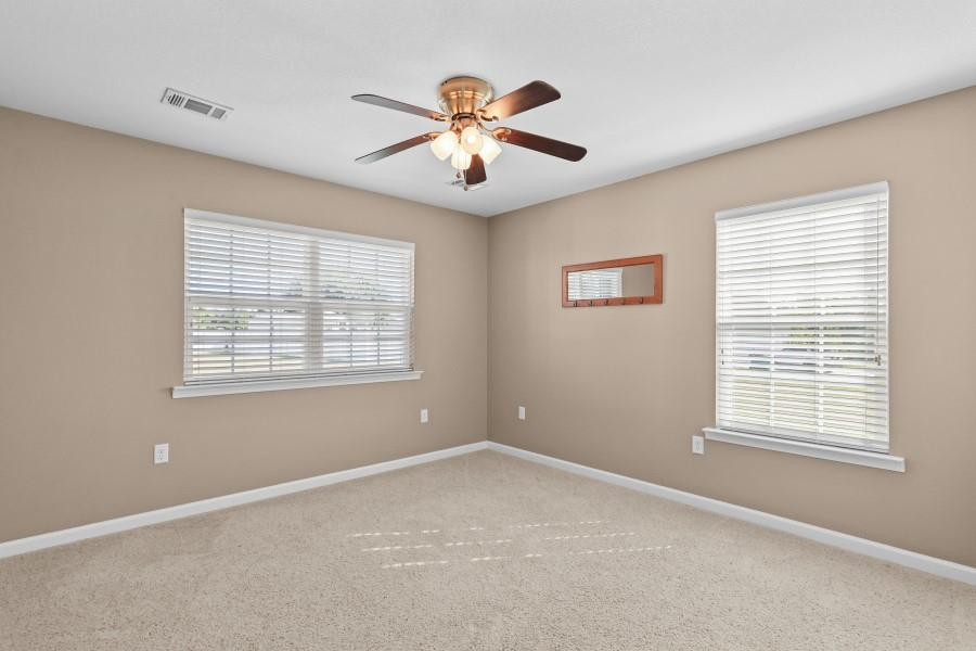 Empty carpeted room with beige walls, two blinded windows, and a ceiling fan with lights.