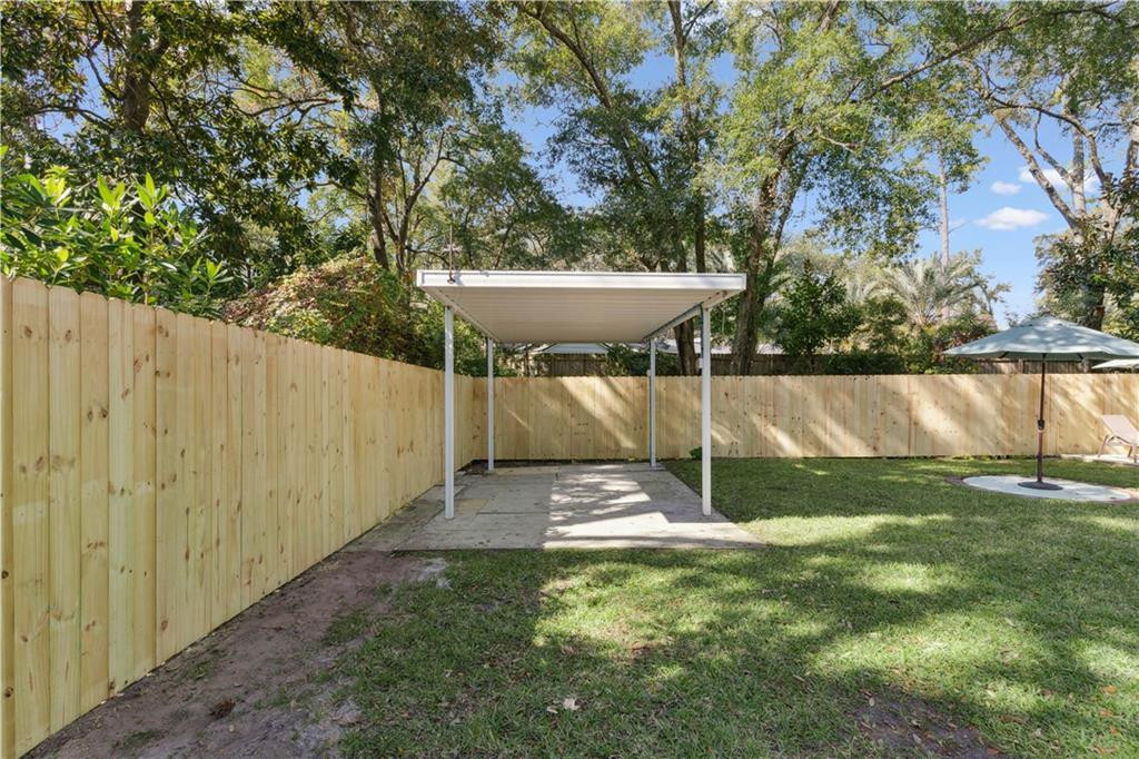 Fenced backyard with a white covered parking shelter, paved pad, lawn, and mature trees.