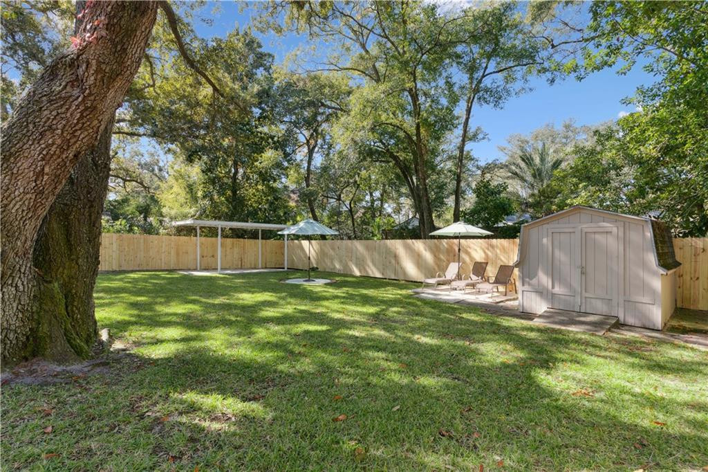 Fenced backyard with a grassy lawn, mature trees, a storage shed, and umbrella-shaded patio seating.