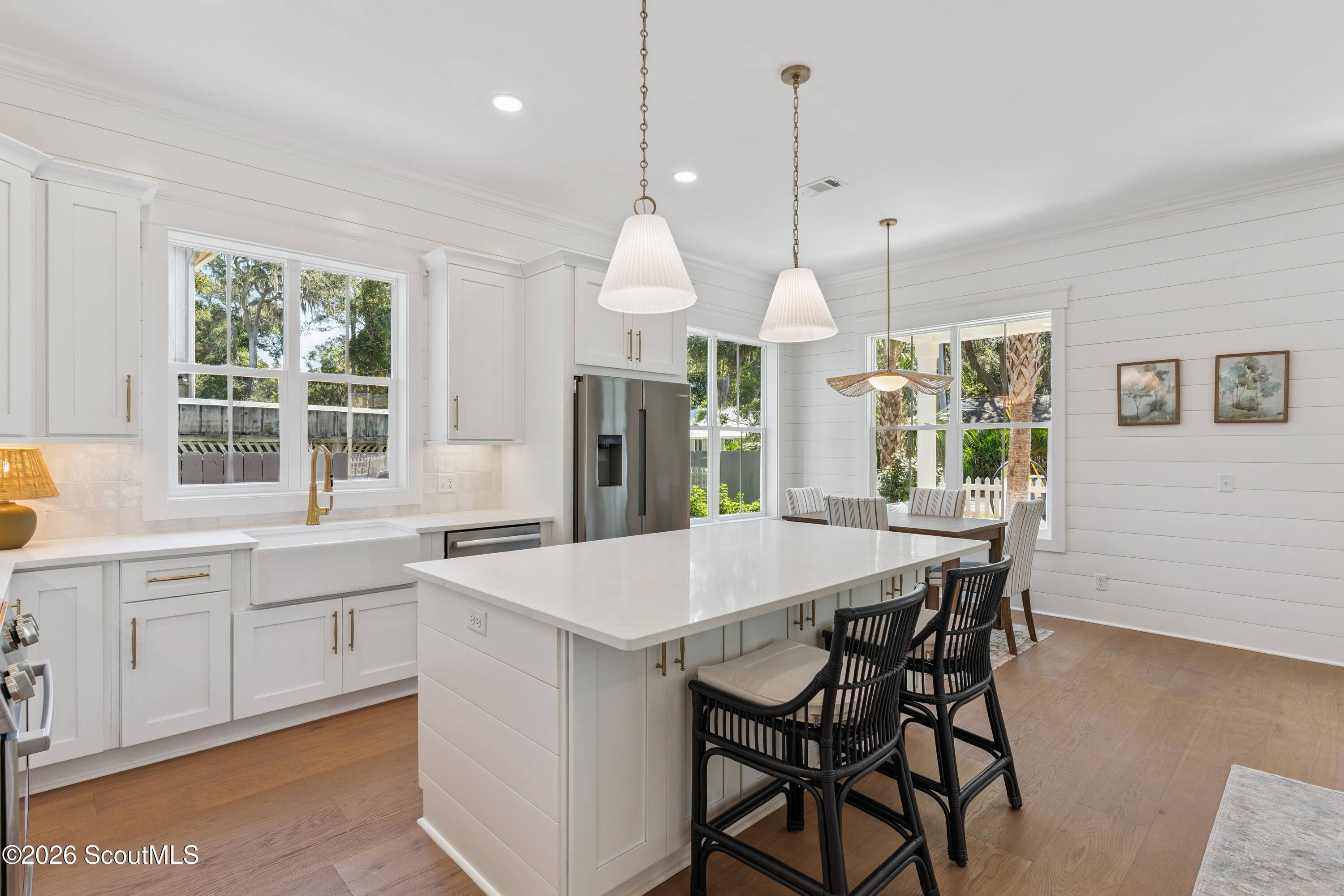 Bright white kitchen with a large island, black bar stools, and windows overlooking trees.