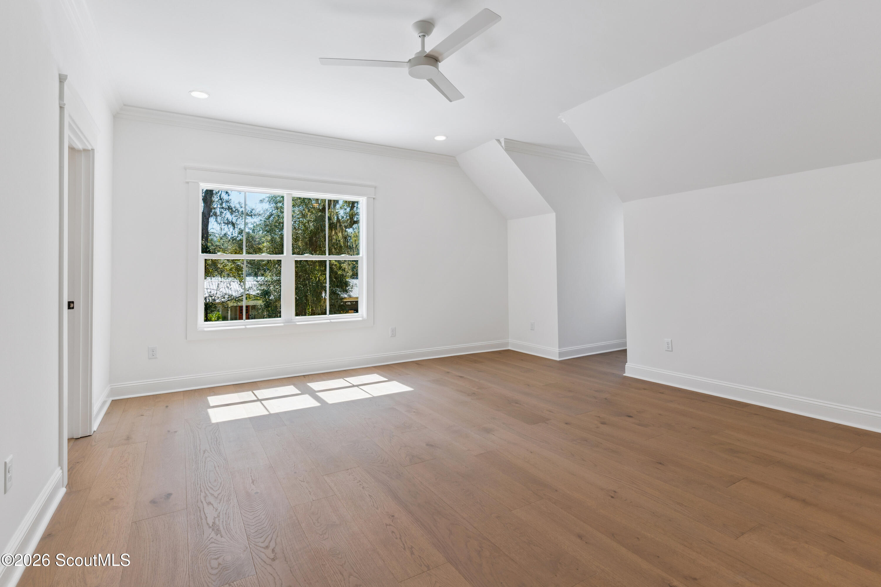 Vacant upstairs room with light wood floors, sloped ceiling, and a large window overlooking trees.