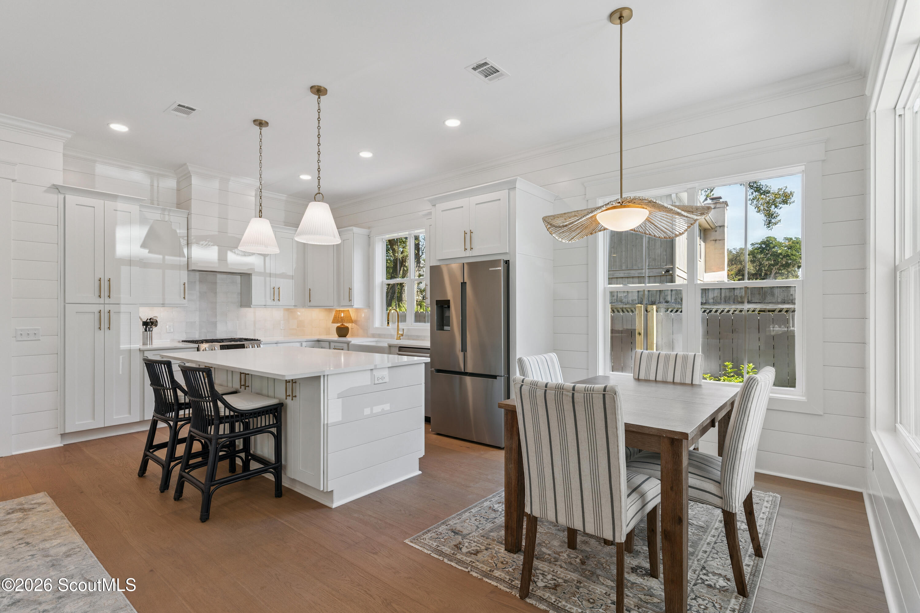 Open-plan kitchen and dining area with a white island, black barstools, stainless steel refrigerator, and large windows.