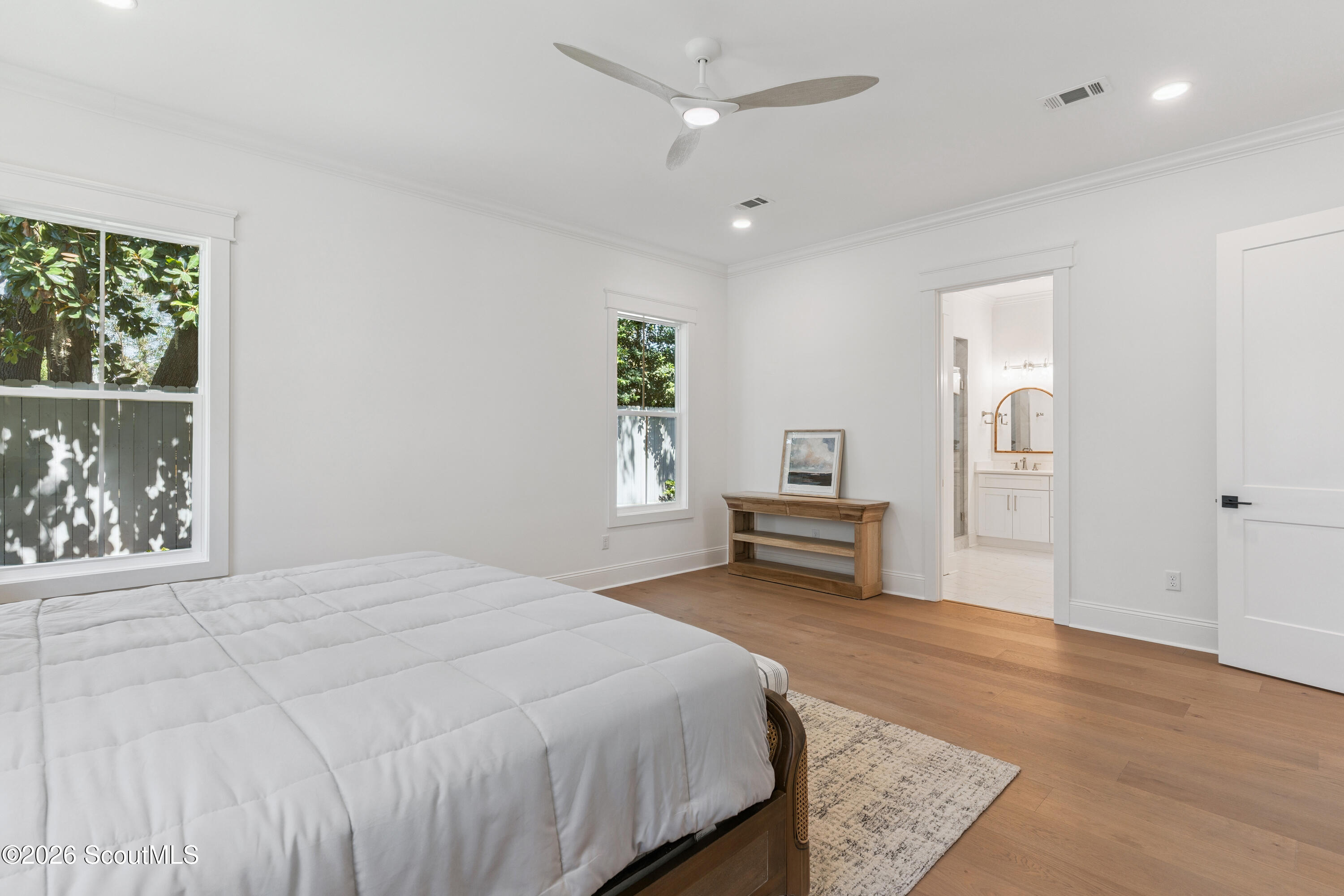 Bright bedroom with a white bed, wood floors, leafy views, and an ensuite bathroom doorway.