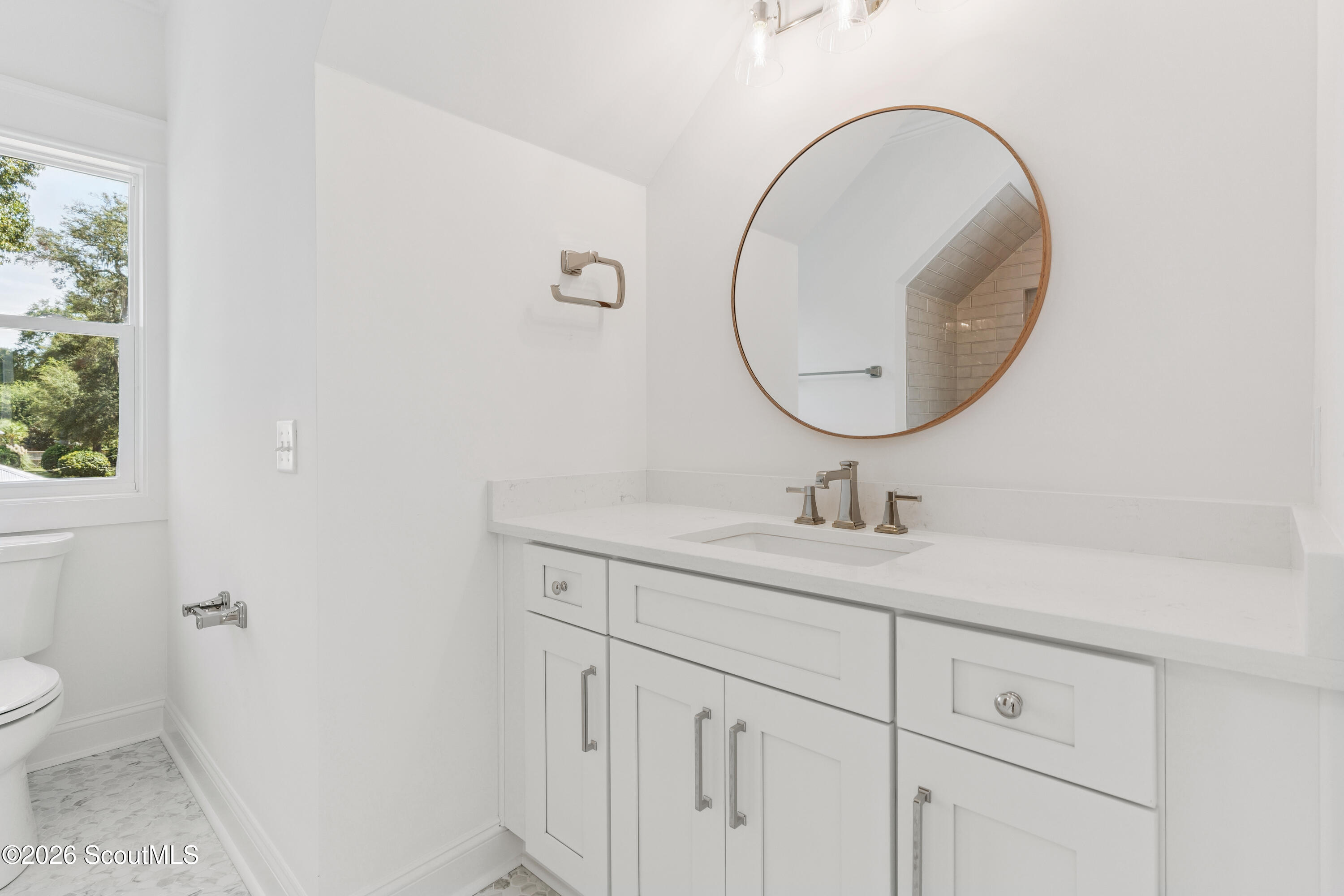 Bright bathroom with a white vanity, round wood-framed mirror, and window.