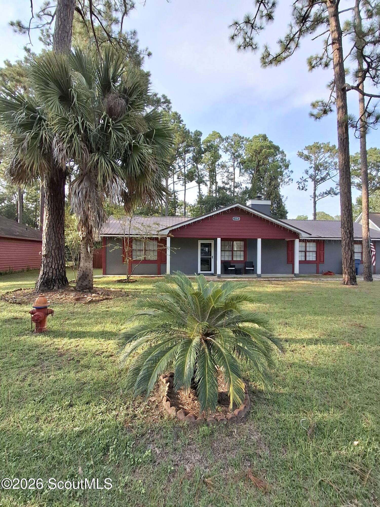Gray single-story house with red trim and a covered entry, framed by palms and pine trees.
