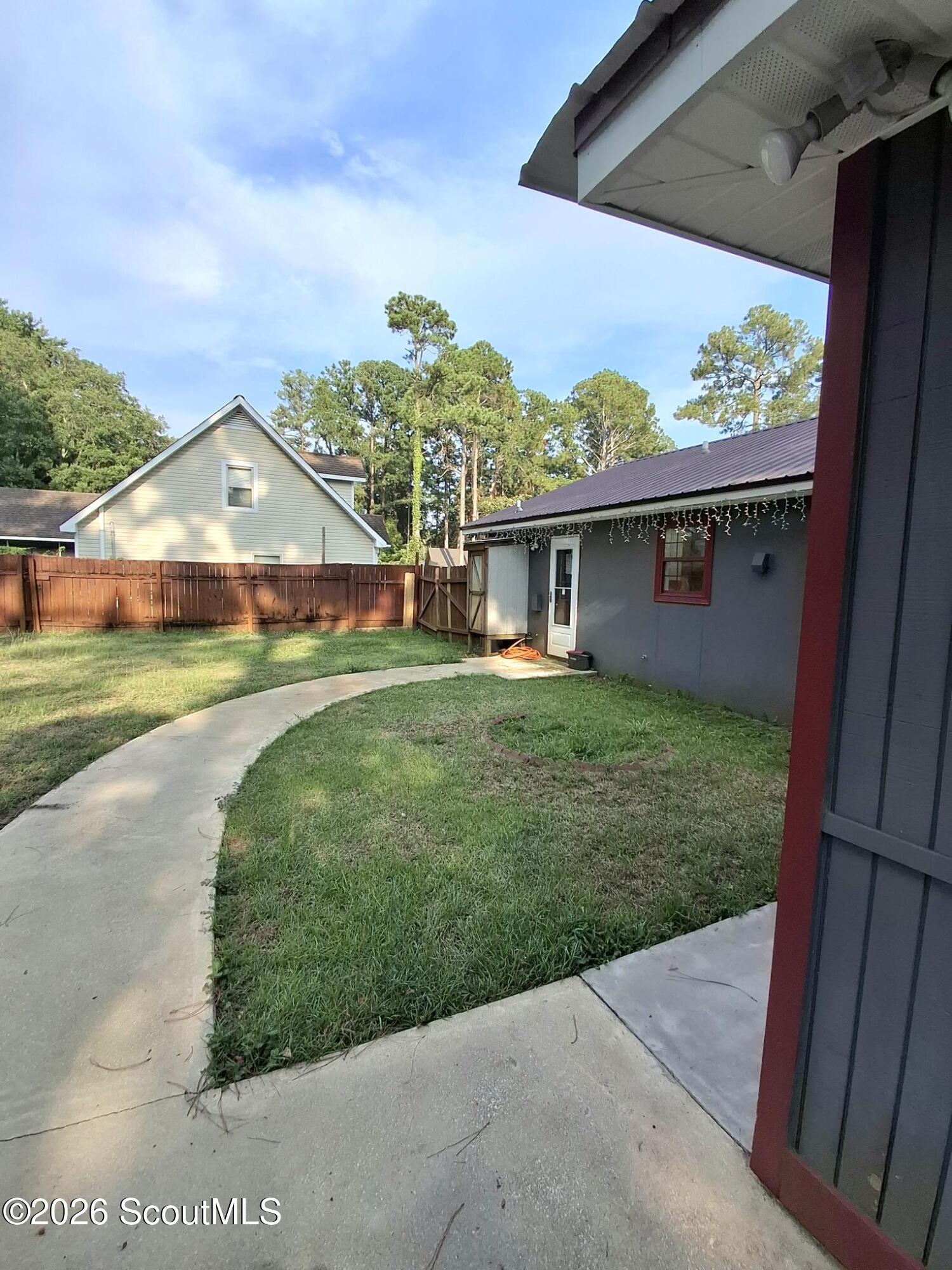 Side yard with a curved concrete walkway, wooden privacy fence, and gray outbuilding with red trim.