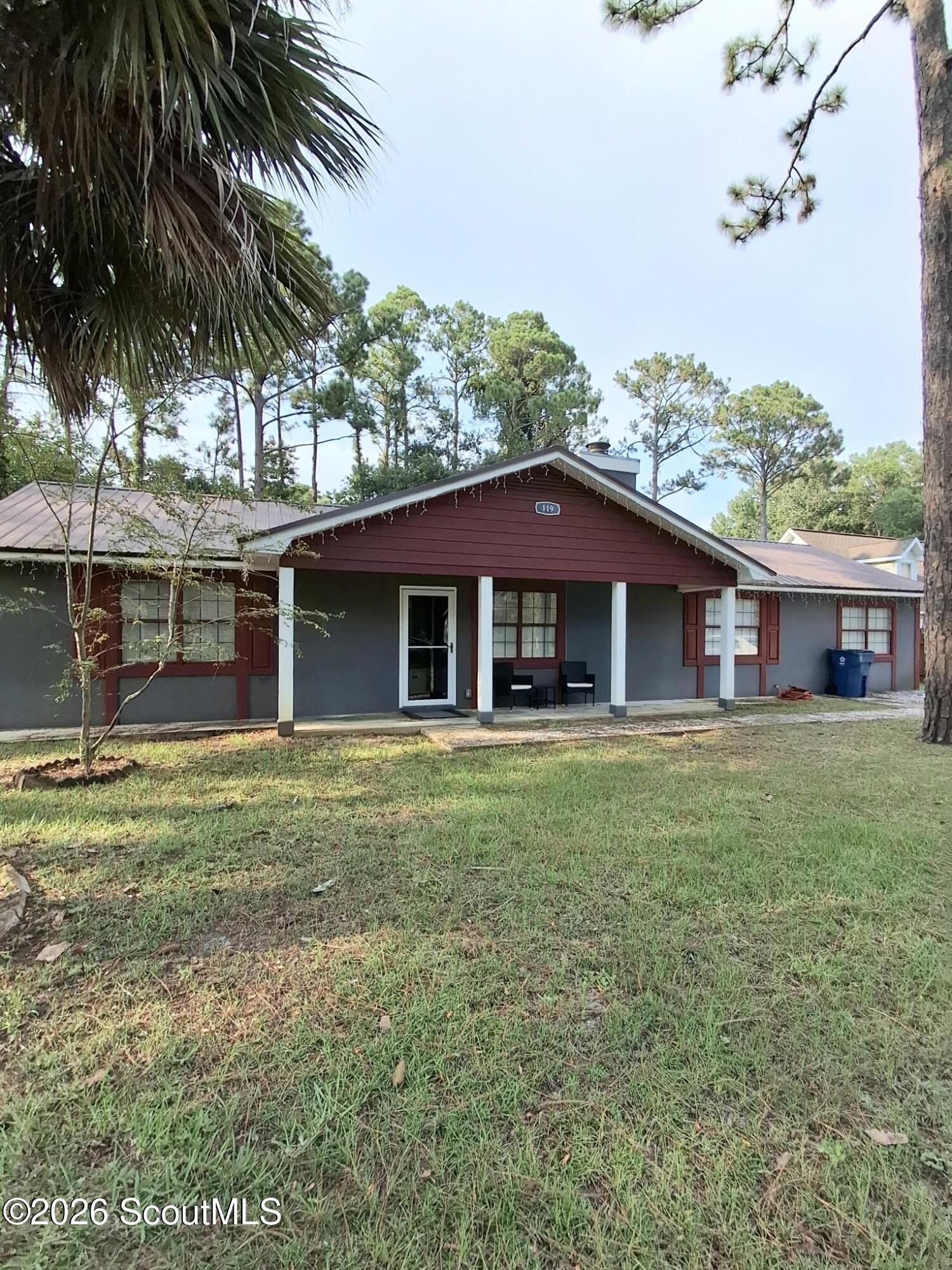 Single-story gray house with burgundy trim, a covered porch, and a grassy yard beneath palm and pine trees.