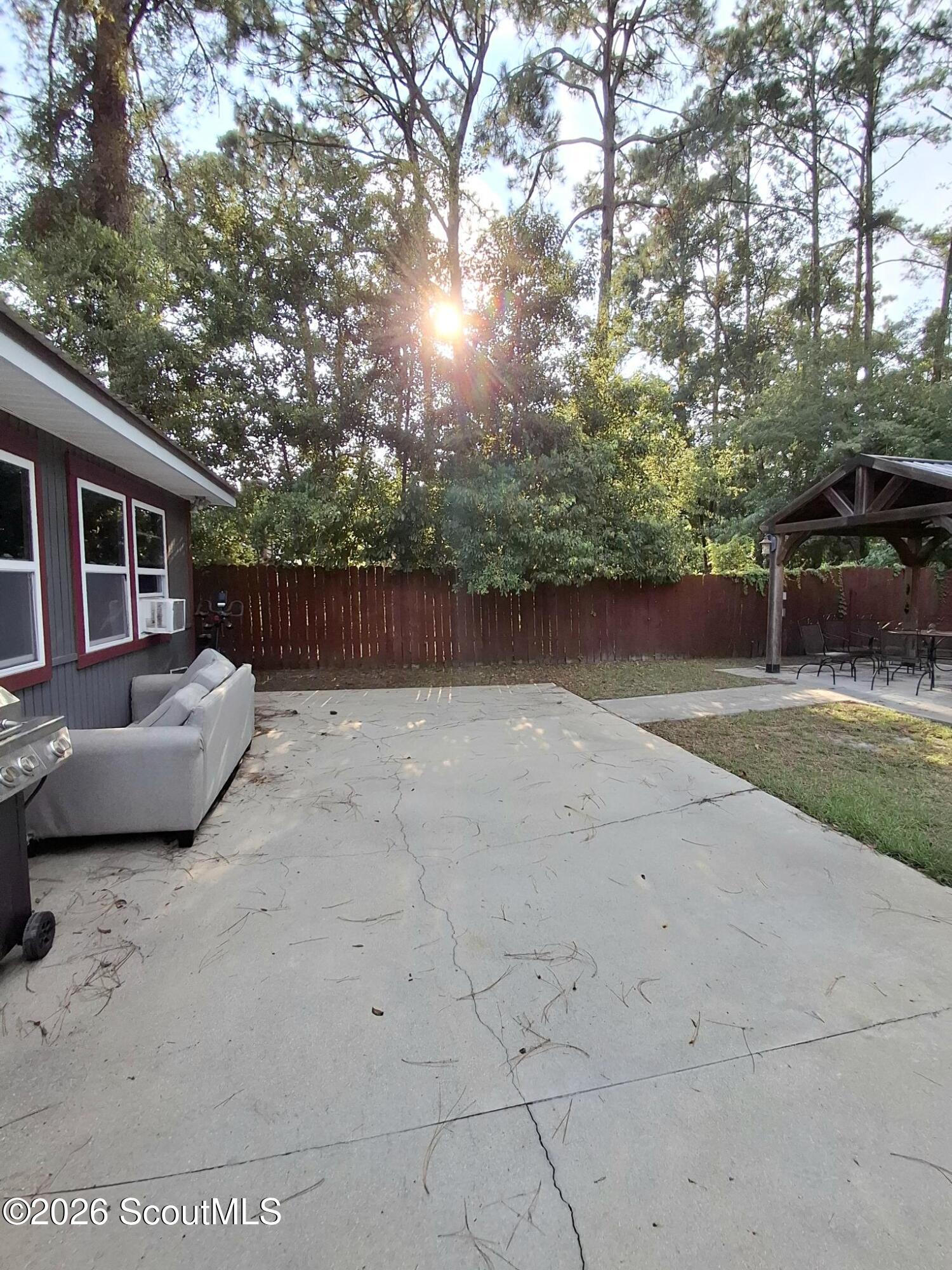 Backyard patio with a concrete slab, outdoor sofa, and covered pergola beneath tall trees.