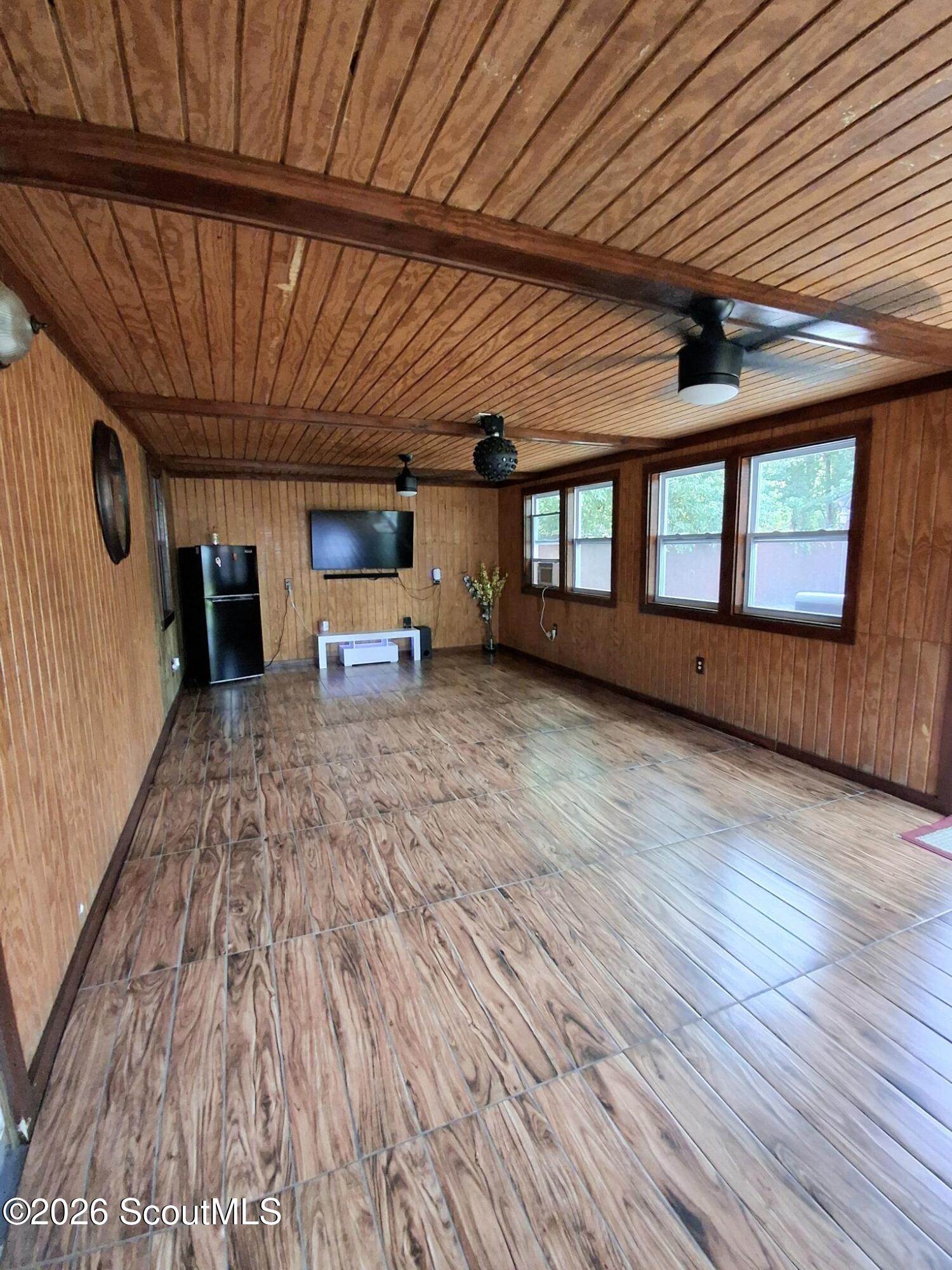 Wood-paneled room with tile flooring, three windows, ceiling fans, a refrigerator, and a wall-mounted television.