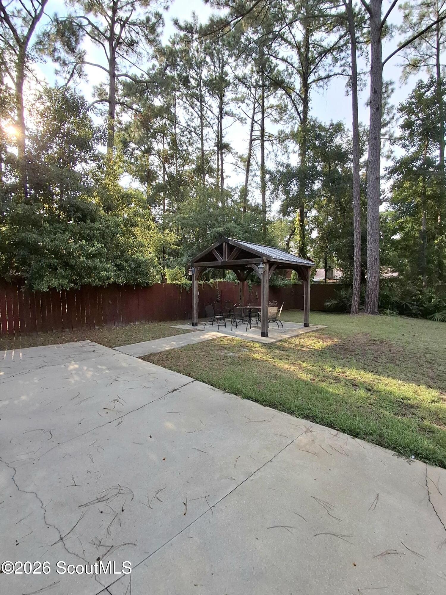Backyard patio with a wooden gazebo, outdoor dining set, and tall trees behind a privacy fence.