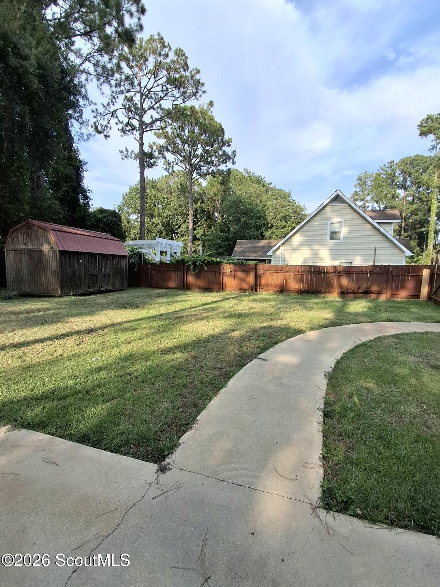 Backyard with a curved concrete walkway, fenced lawn, and a wood shed with a red roof.