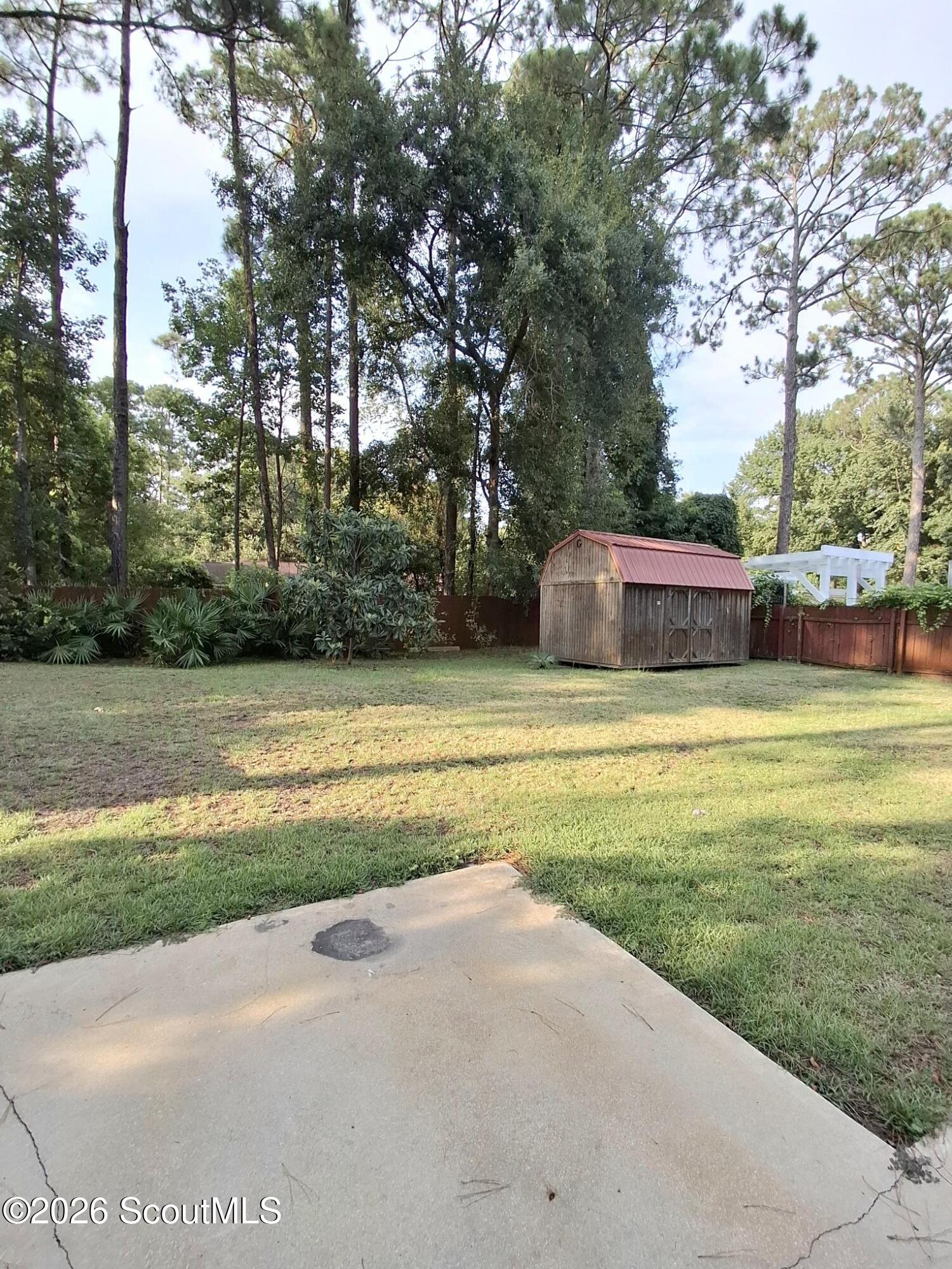 Fenced backyard with a grassy lawn, red-roofed storage shed, mature trees, and concrete patio.