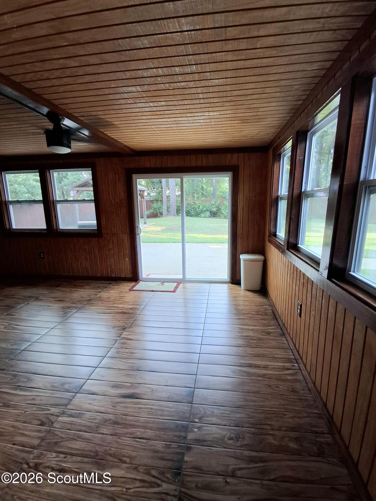 Empty wood-paneled sunroom with tiled floors, a ceiling fan, and windows overlooking the backyard.