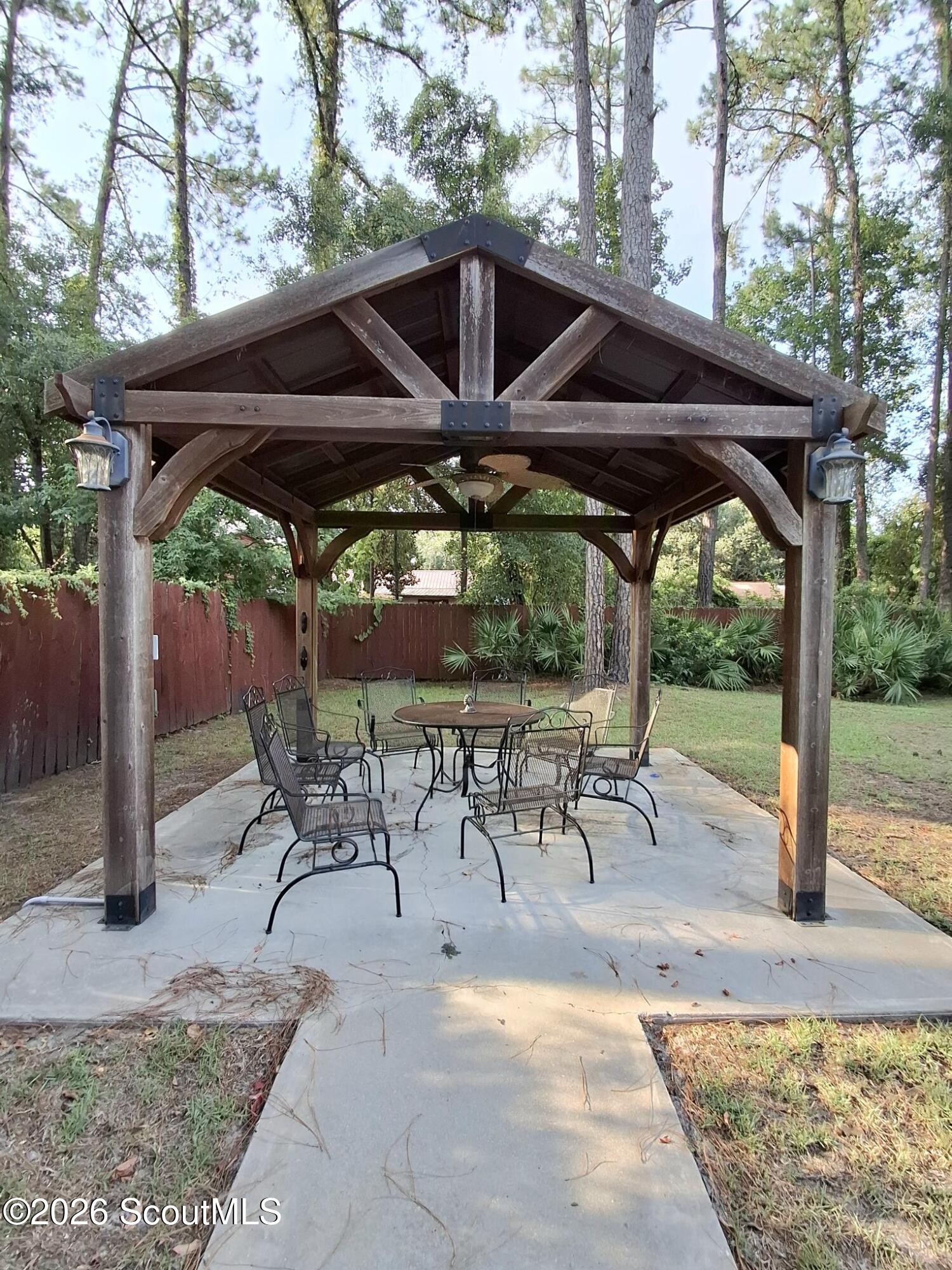Wooden backyard gazebo with a peaked roof, round table, metal chairs, and paved patio.