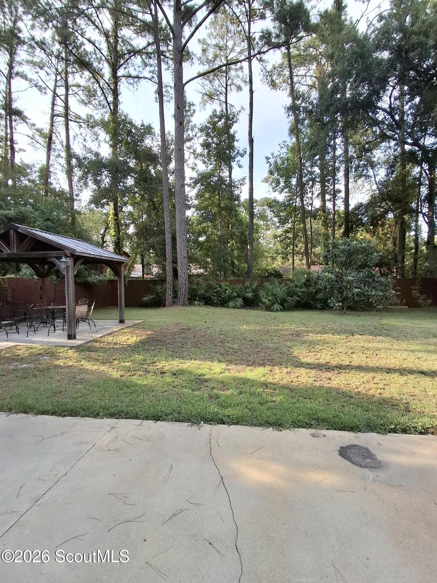 Tree-shaded backyard with a covered pavilion, outdoor dining set, and broad concrete patio.