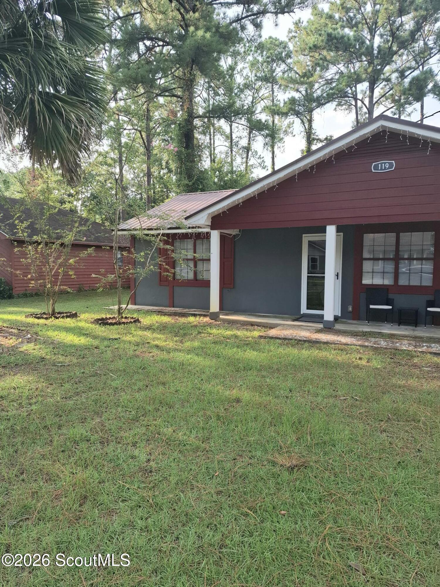 Home with dark red siding and a covered porch, set behind a grassy lawn and mature trees.
