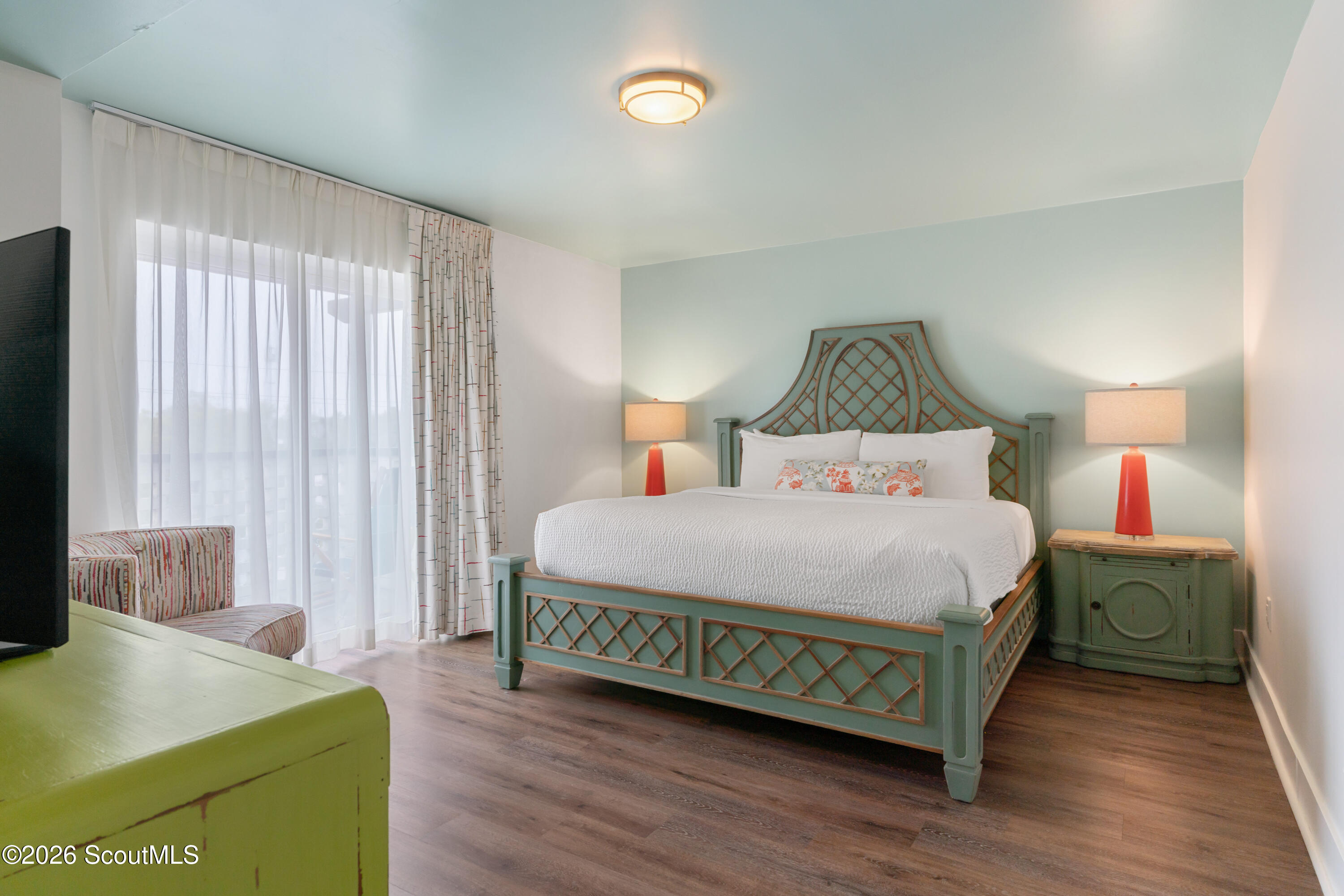 Bedroom with a decorative teal-green bed, white bedding, and red bedside lamps beside sheer-curtained windows.