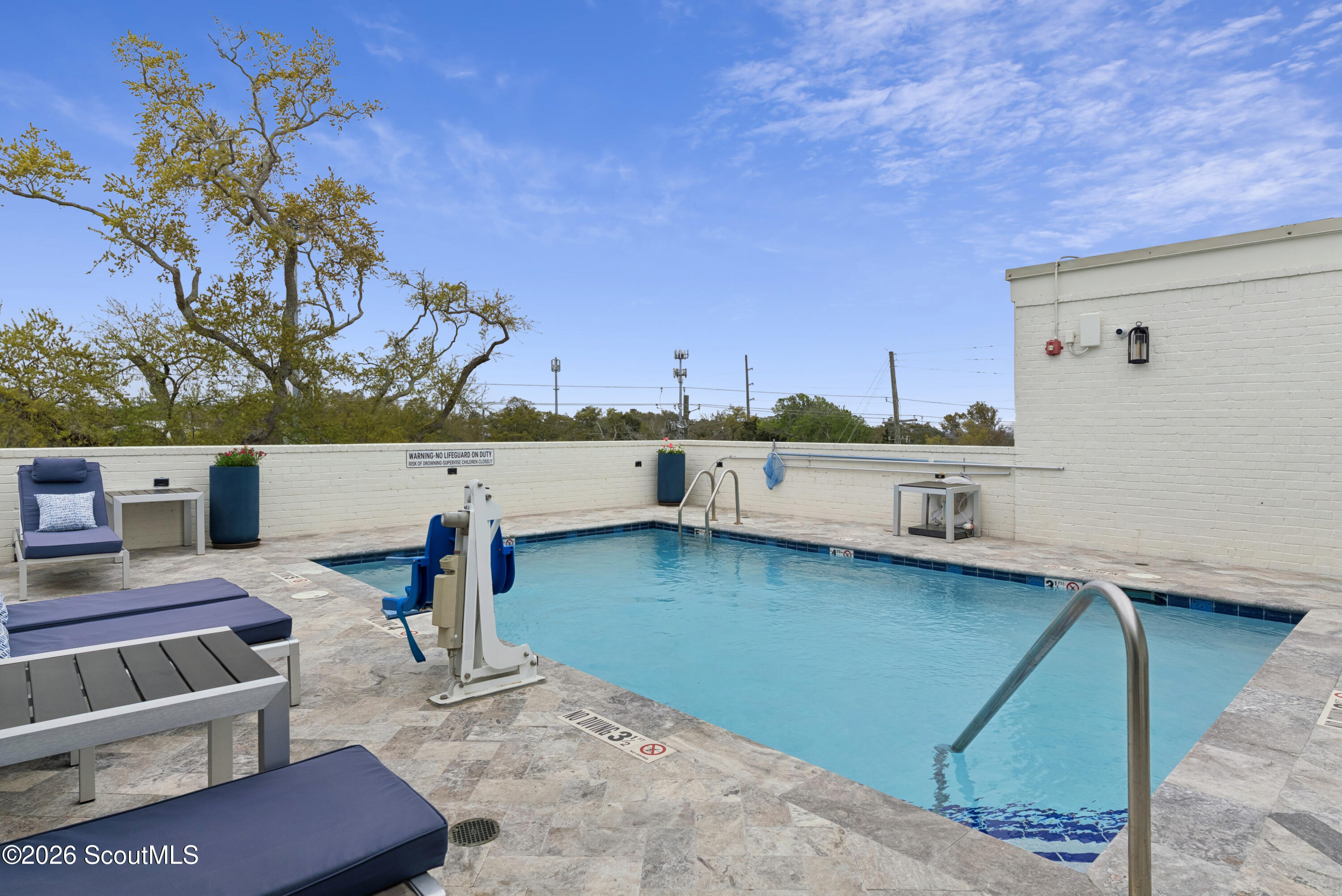 Outdoor swimming pool with an accessible lift, metal ladder, and cushioned loungers on a stone deck.