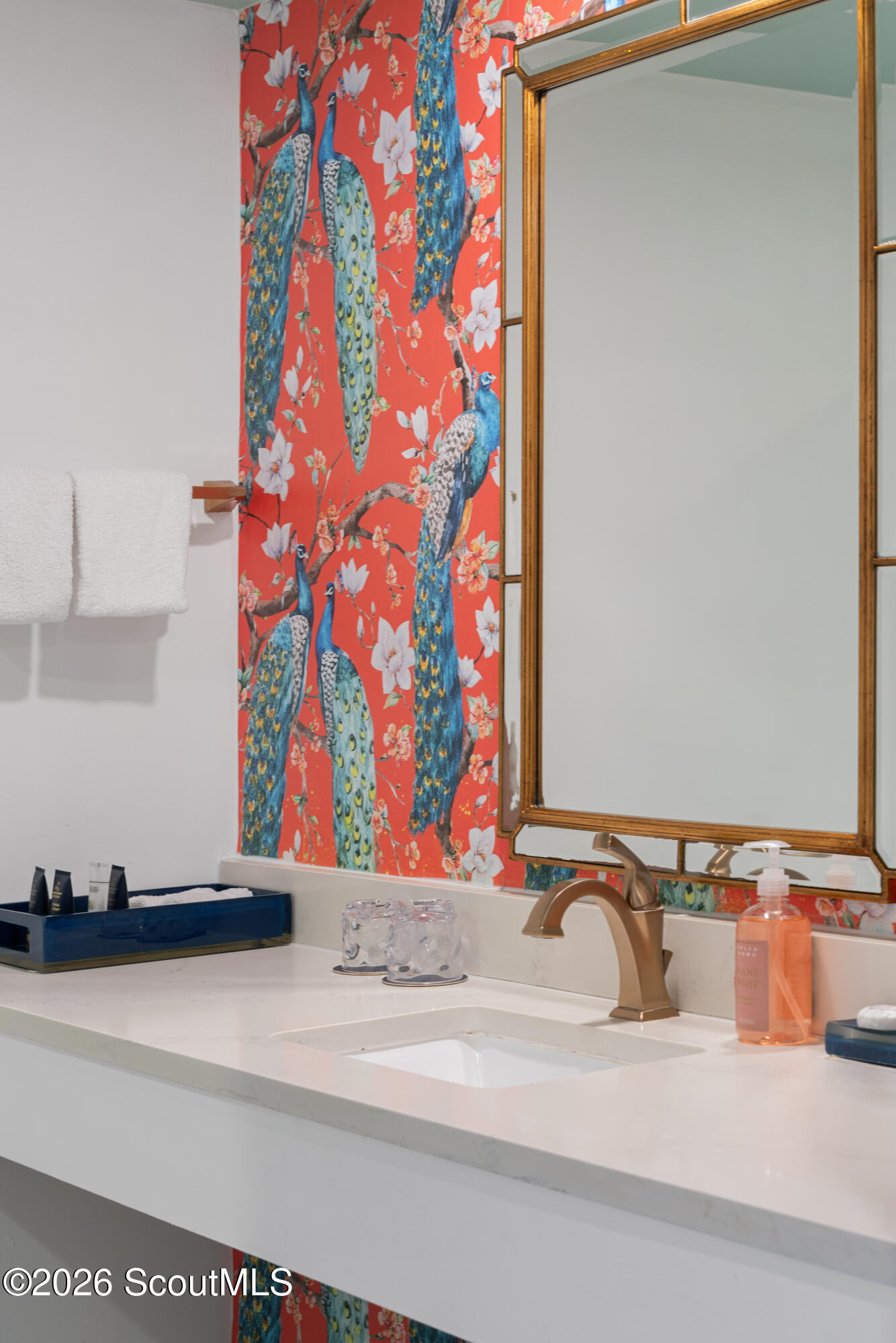 Bathroom vanity with a bronze faucet and mirror beside vivid peacock-and-floral wallpaper.