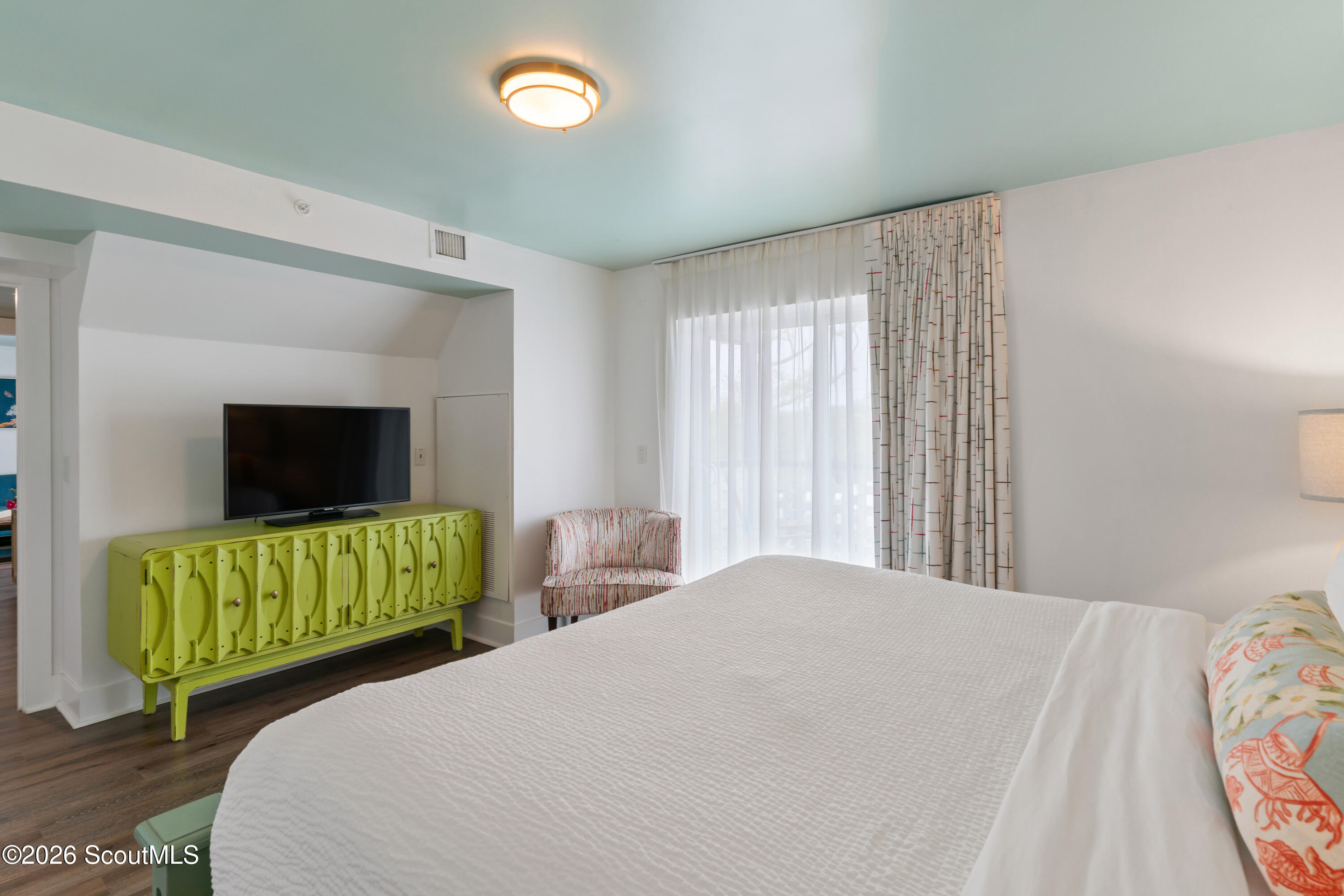 Bedroom with a white bed, lime-green cabinet and television, patterned chair, and sheer-curtained window.