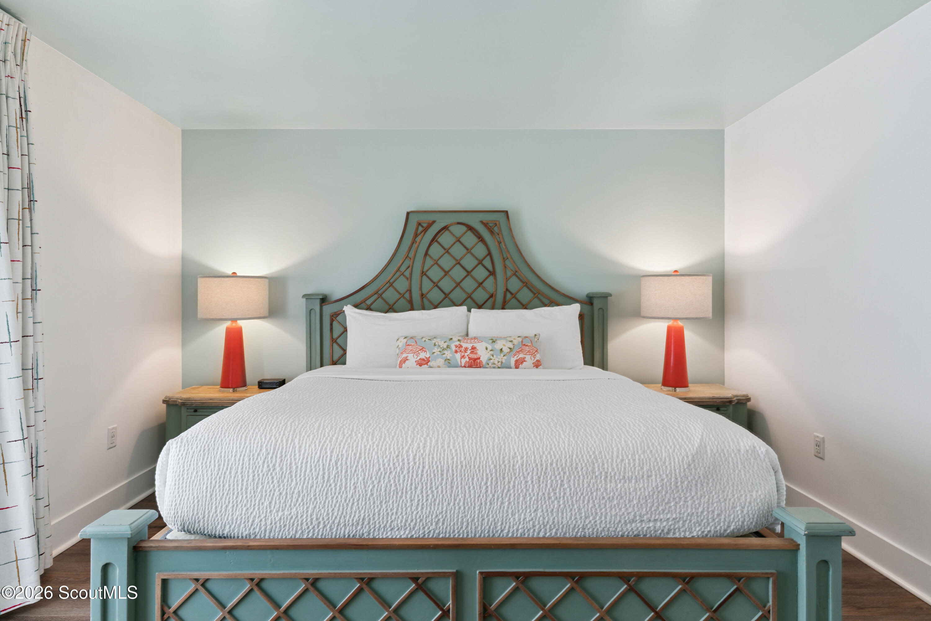 Bedroom with a white-quilted bed, ornate blue-green headboard, matching red lamps, and patterned curtains.