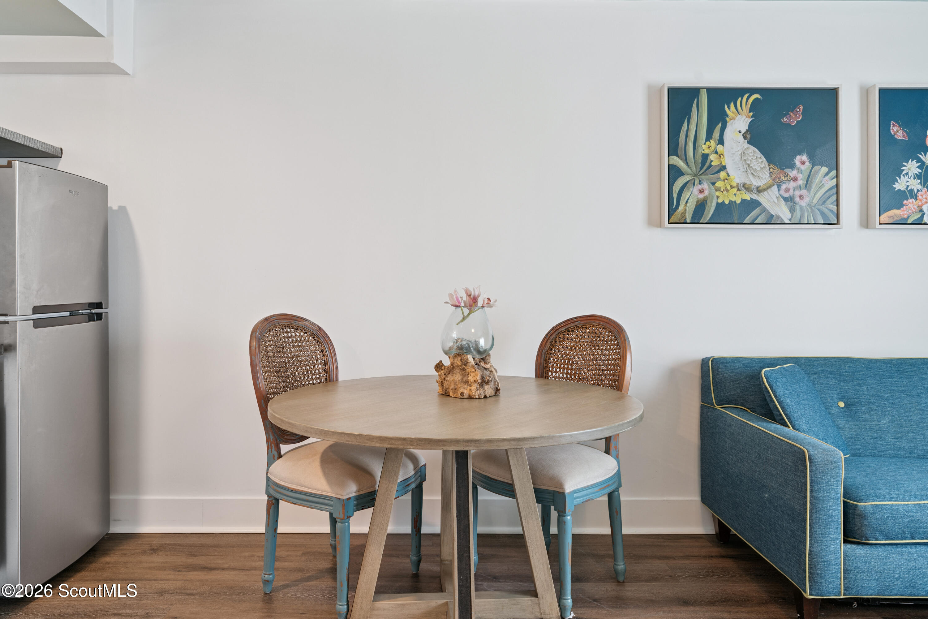 Dining area with a round table and cane-back chairs beside a blue sofa and stainless-steel refrigerator.