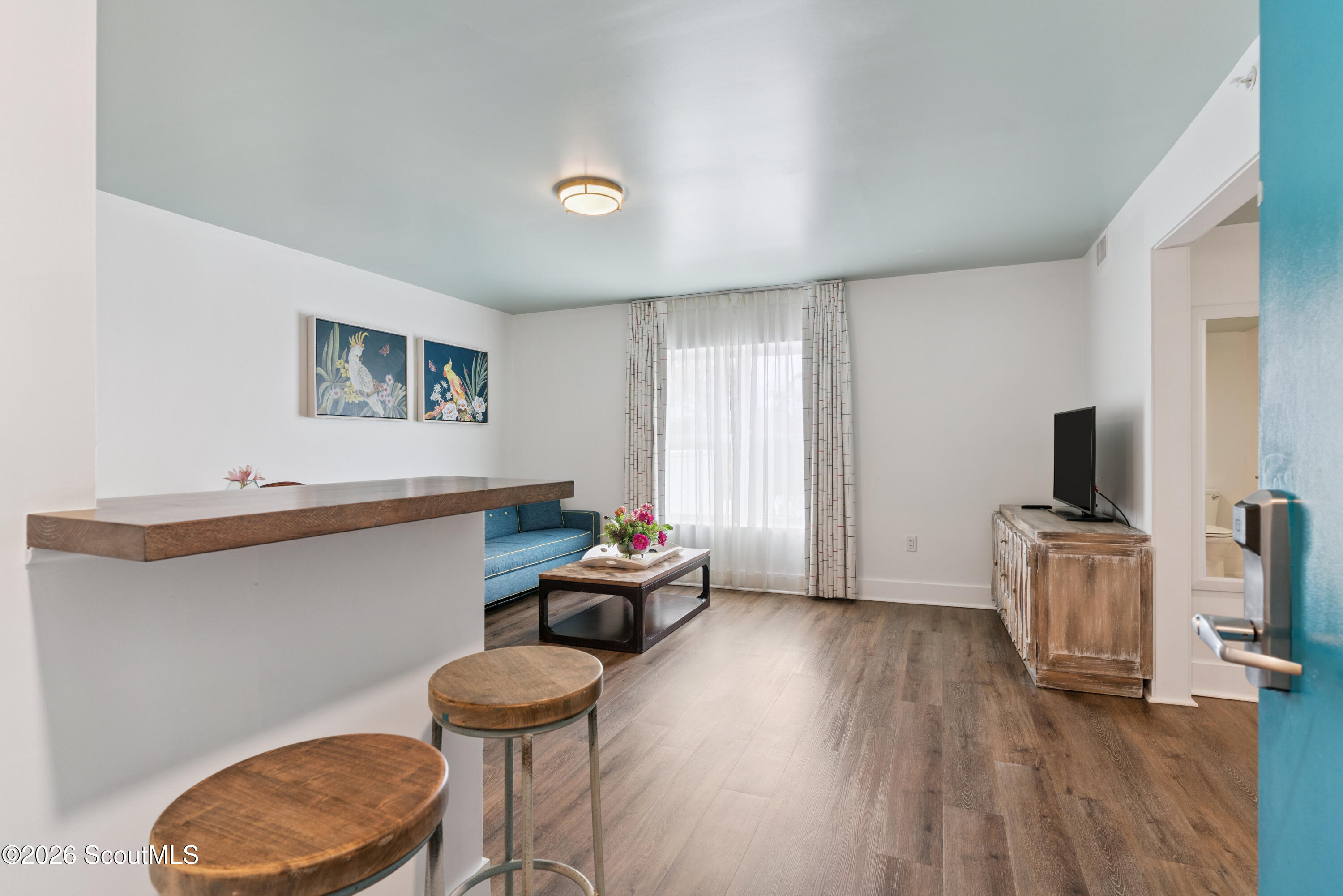 Condominium living room with blue sofa, wooden breakfast bar, and bright curtained window.