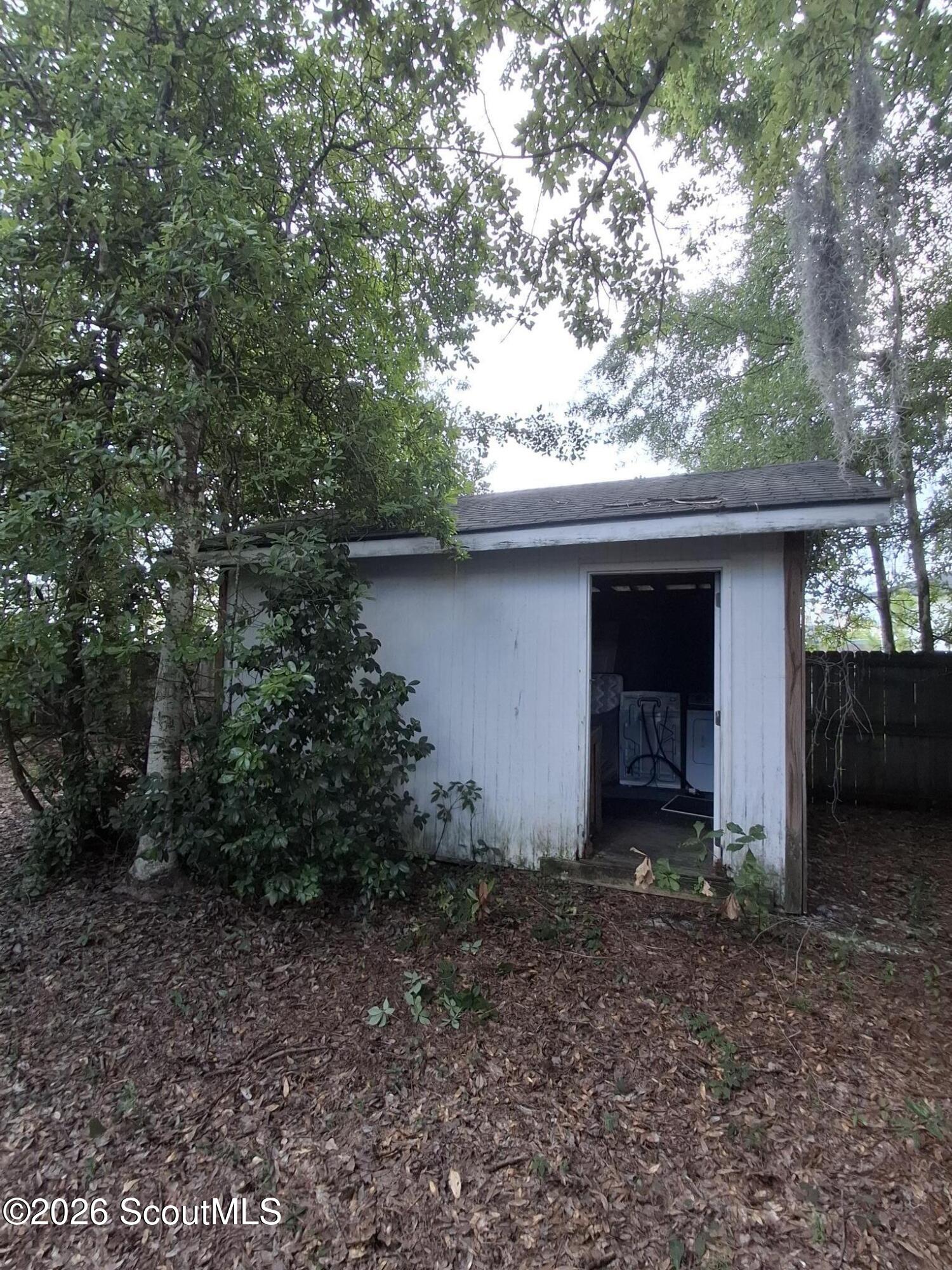 White outbuilding with an open doorway among trees, with a washing machine visible inside.