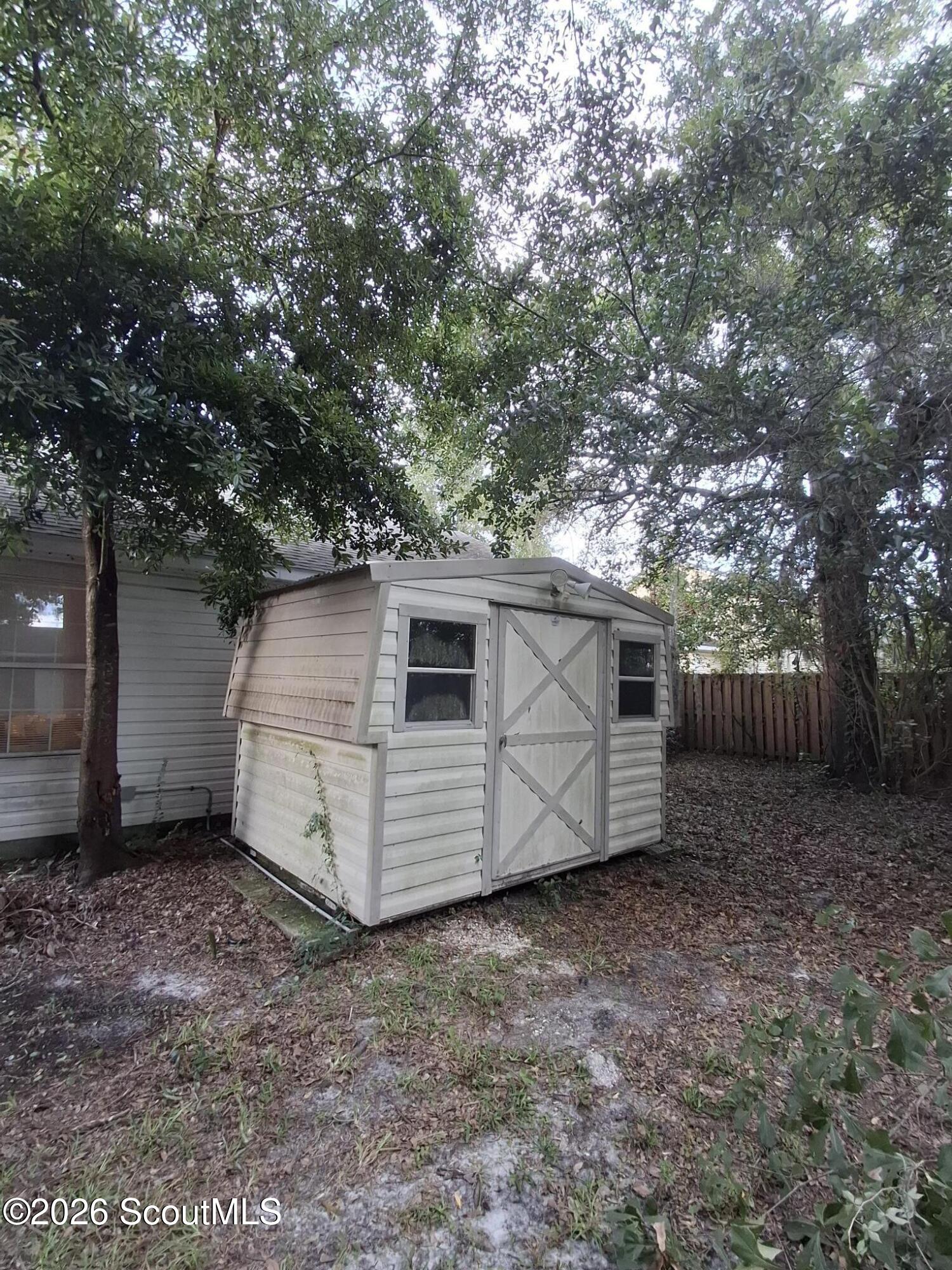 White backyard storage shed with cross-braced doors beneath a dense tree canopy.