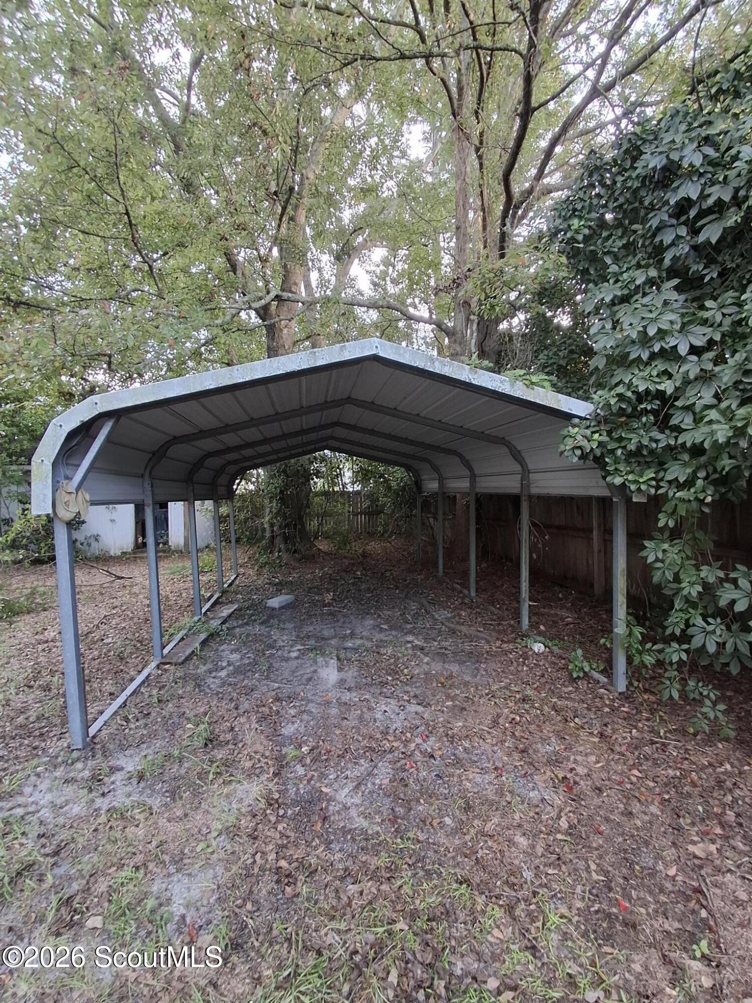 Metal-roofed carport beneath mature trees in a wooded backyard.
