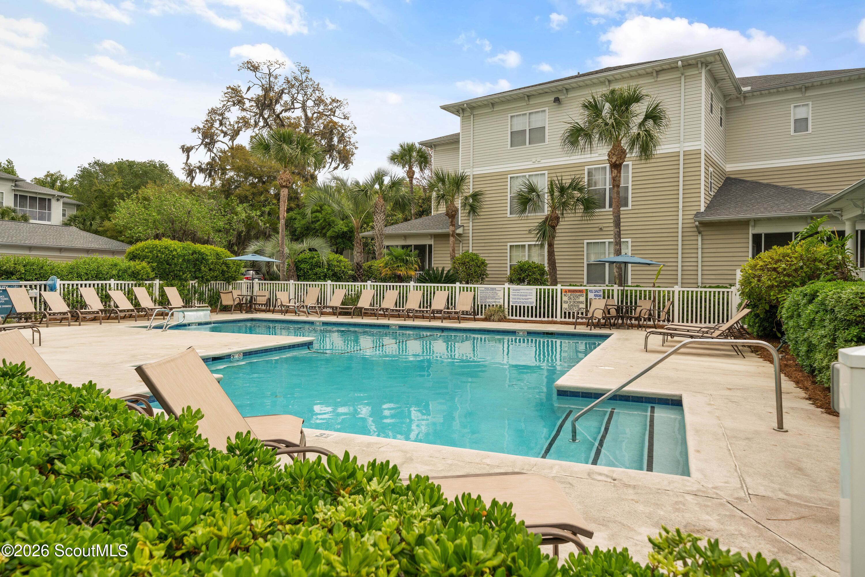 Outdoor pool beside a beige multi-story building, with lounge chairs, palm trees, and a shallow entry.