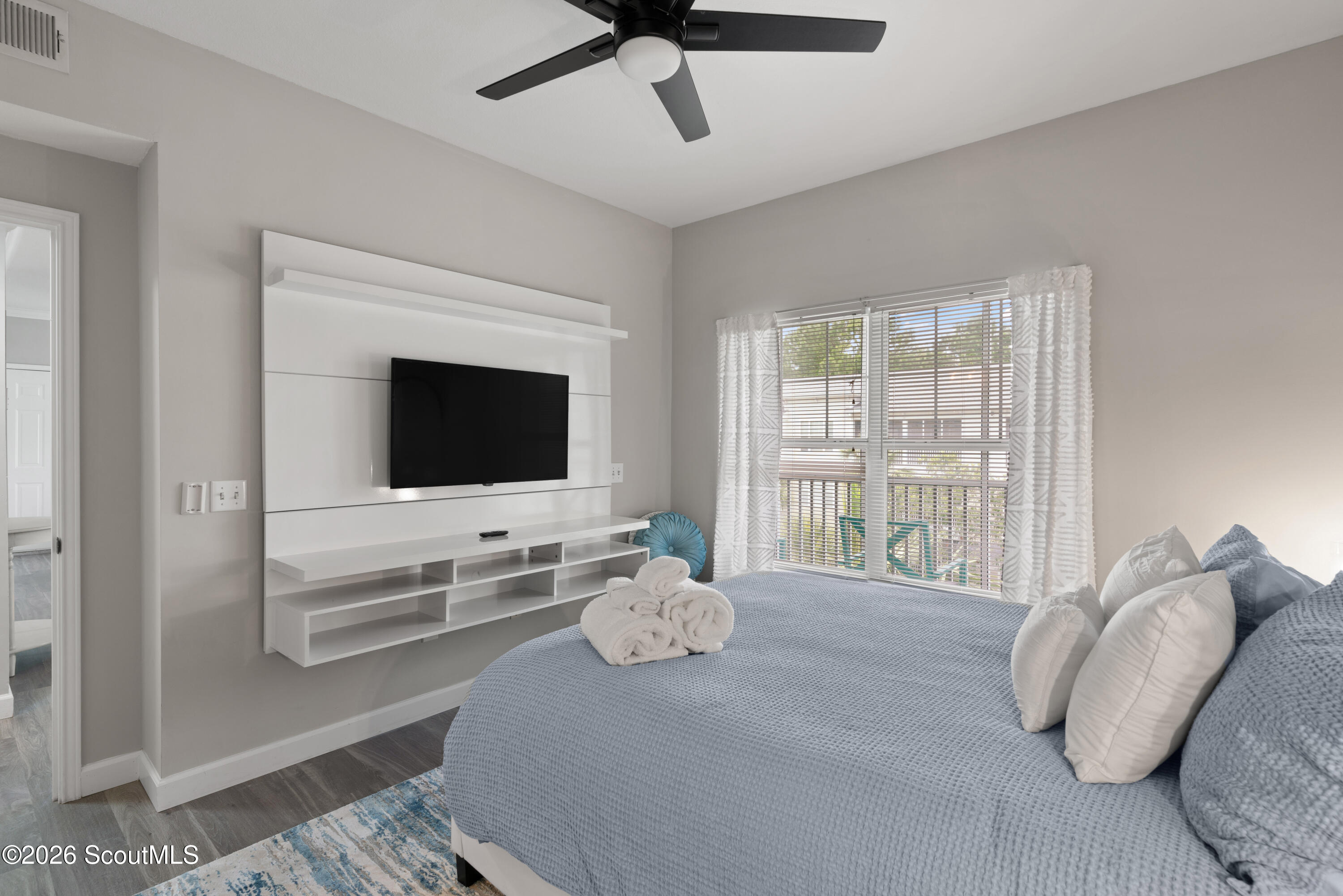 Bedroom with blue-gray bedding, a wall-mounted TV, and a bright window overlooking a deck.
