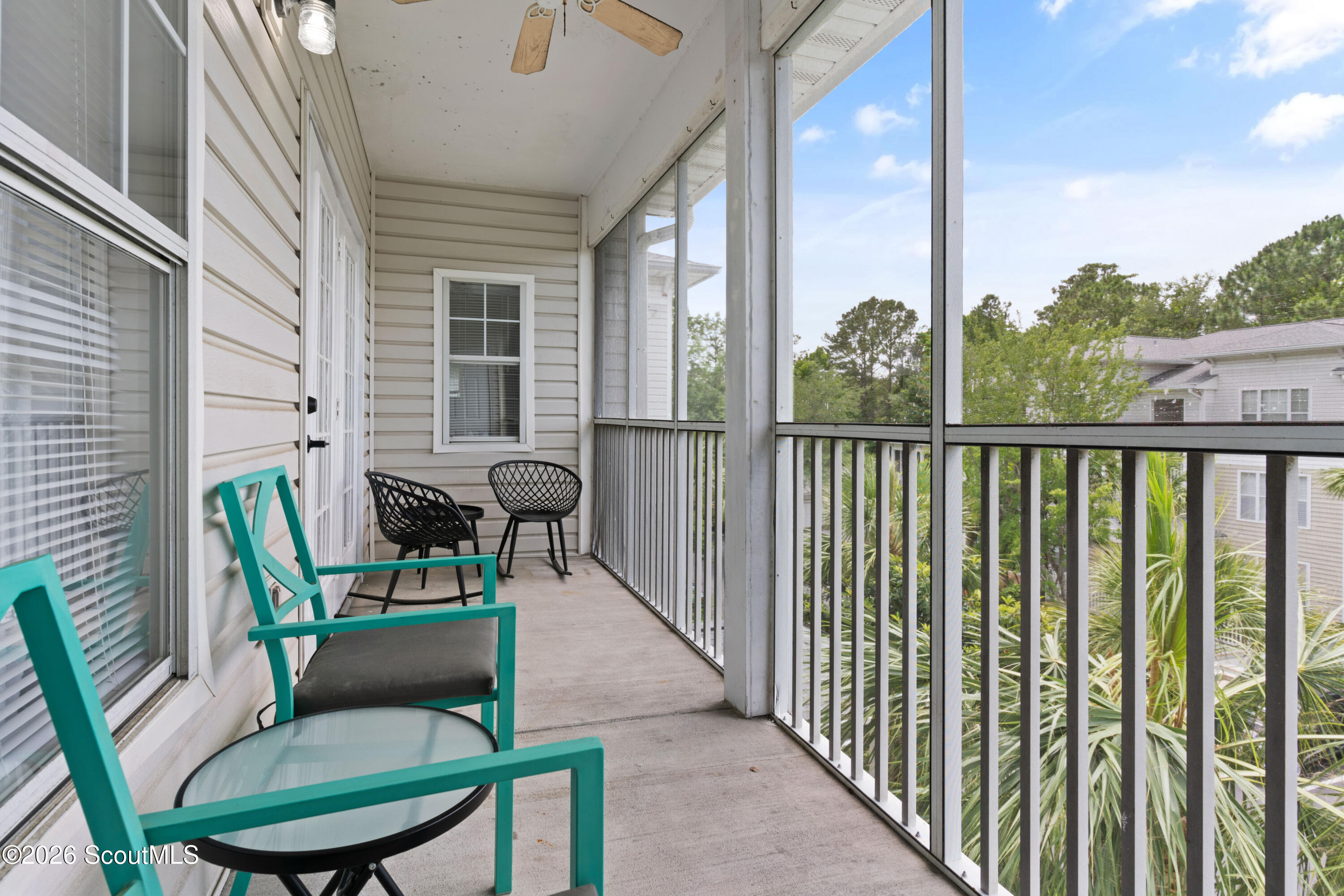 Screened balcony furnished with turquoise chairs and a glass-topped table, overlooking greenery and a neighboring building.