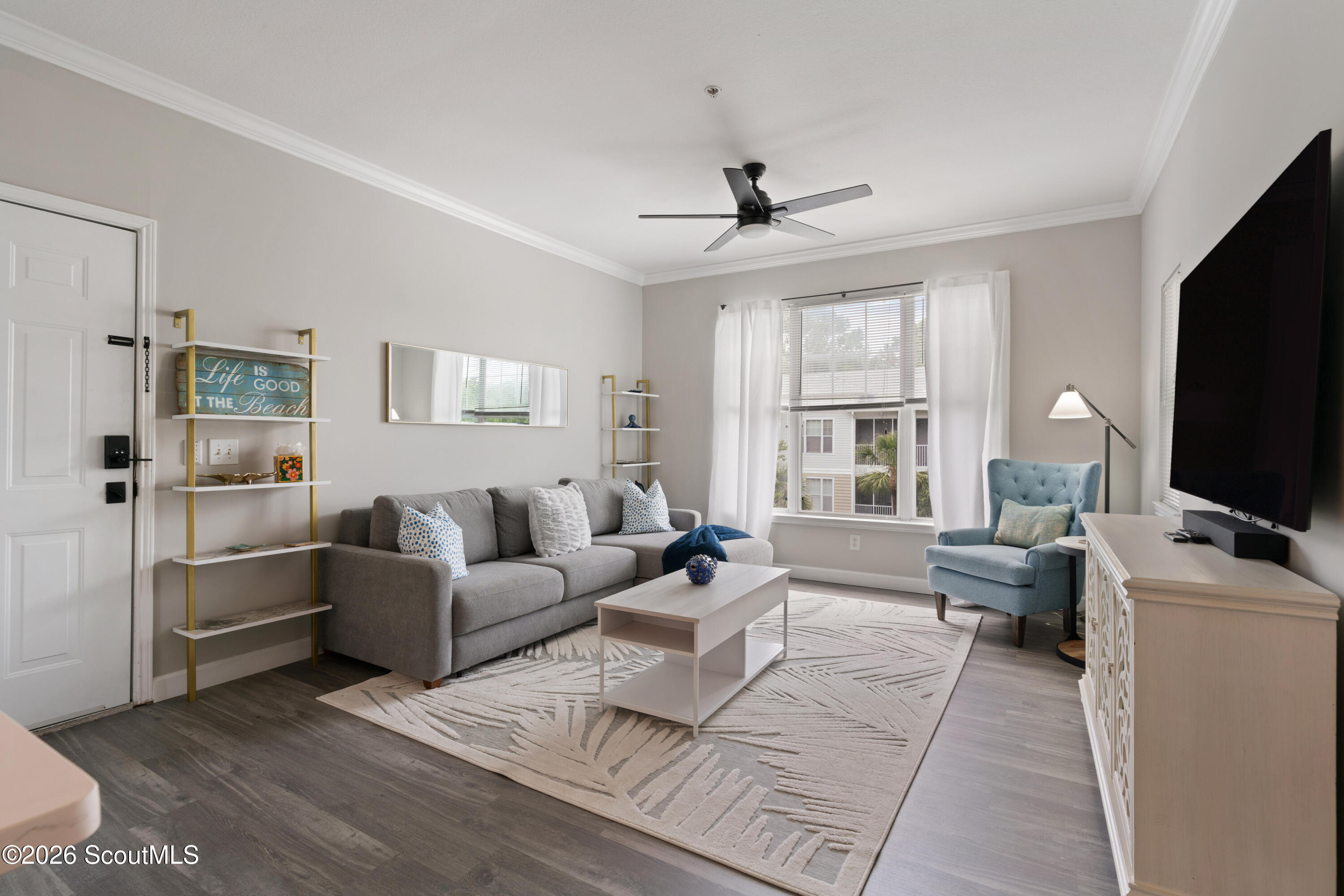 Staged living room with a gray sofa, blue armchair, patterned rug, and large curtained window.
