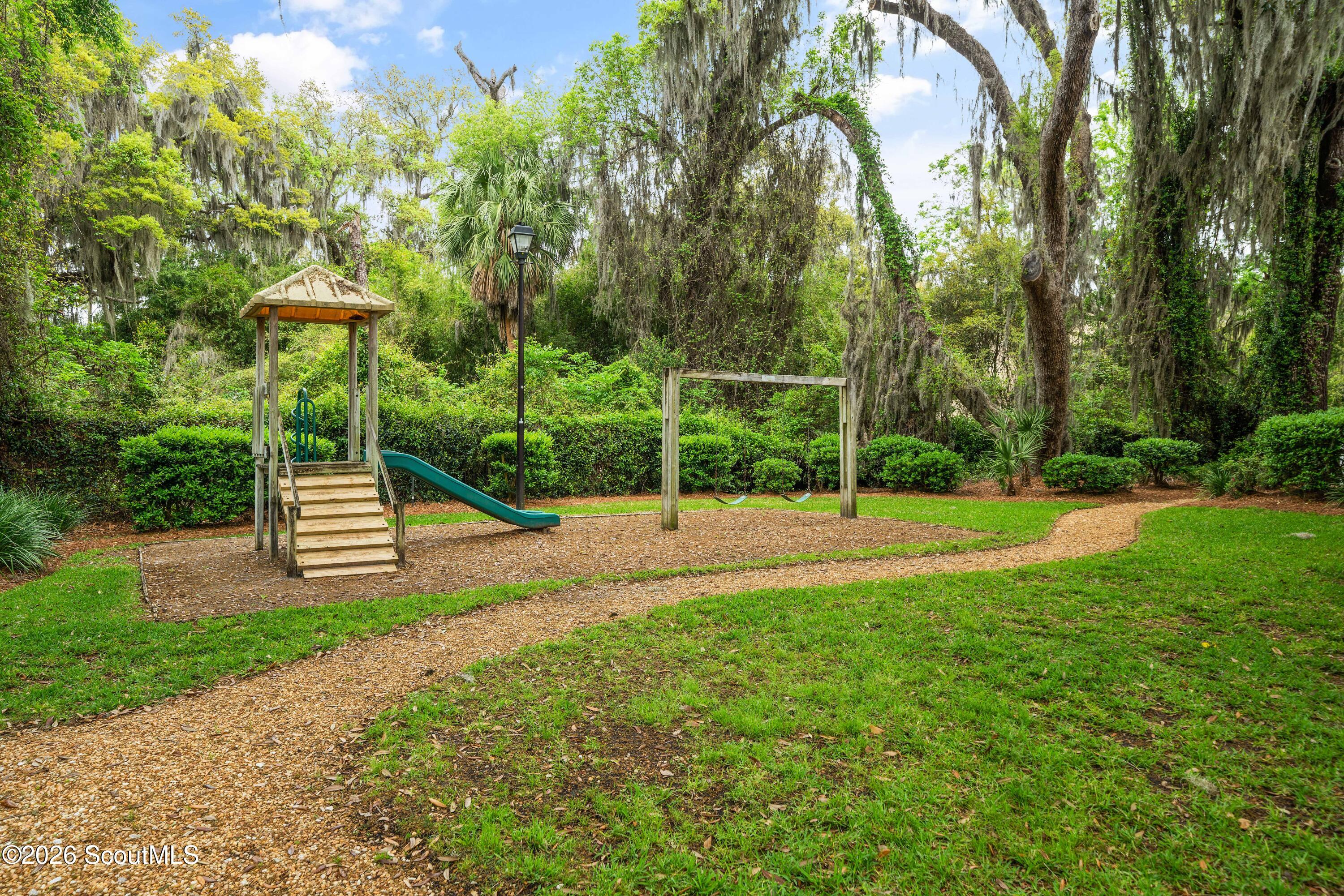 Playground with a wooden play tower, green slide, swings, and a winding path beneath moss-draped trees.
