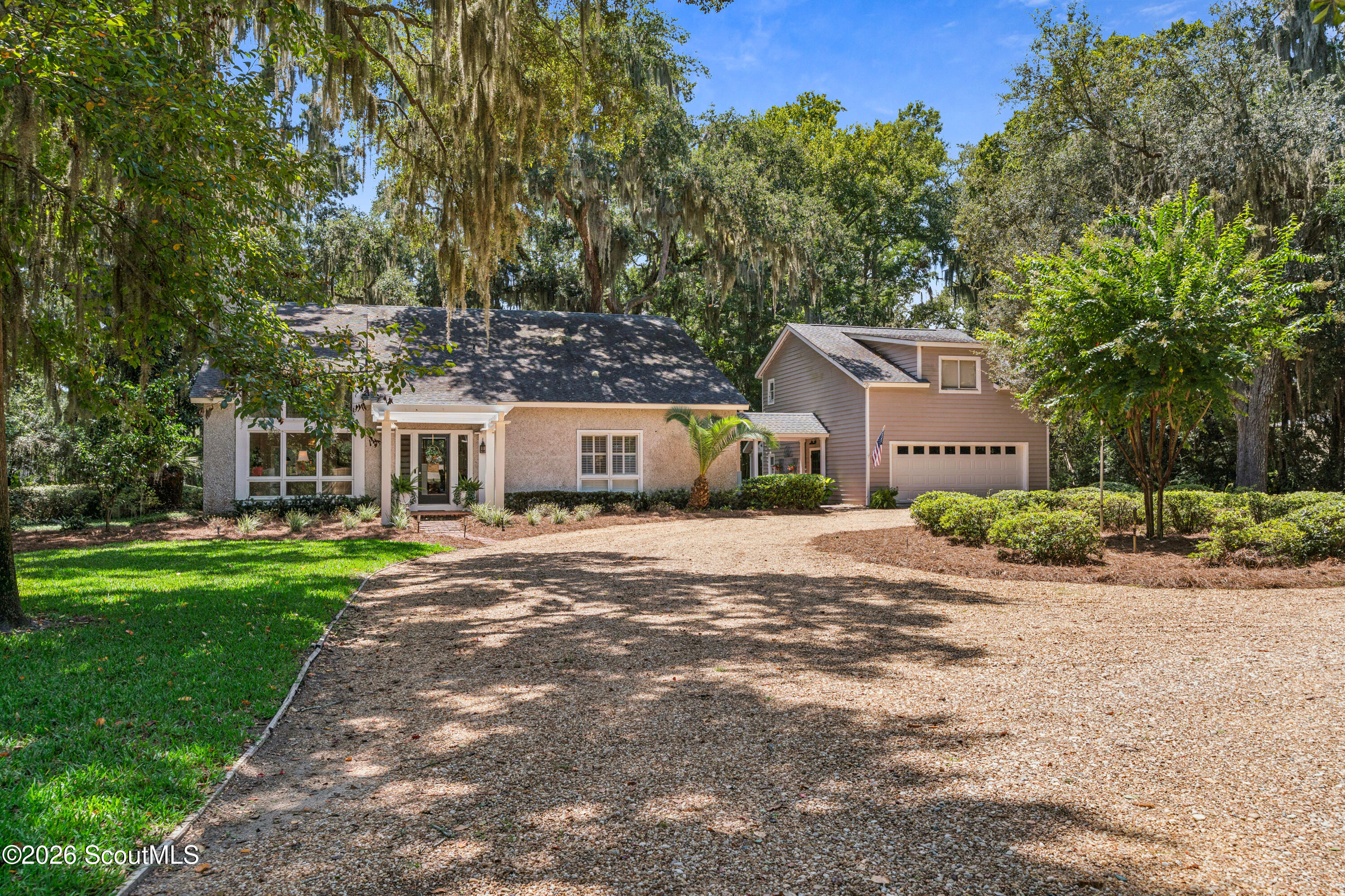 Beige home with an attached two-story garage, broad gravel driveway, and mature moss-draped trees.