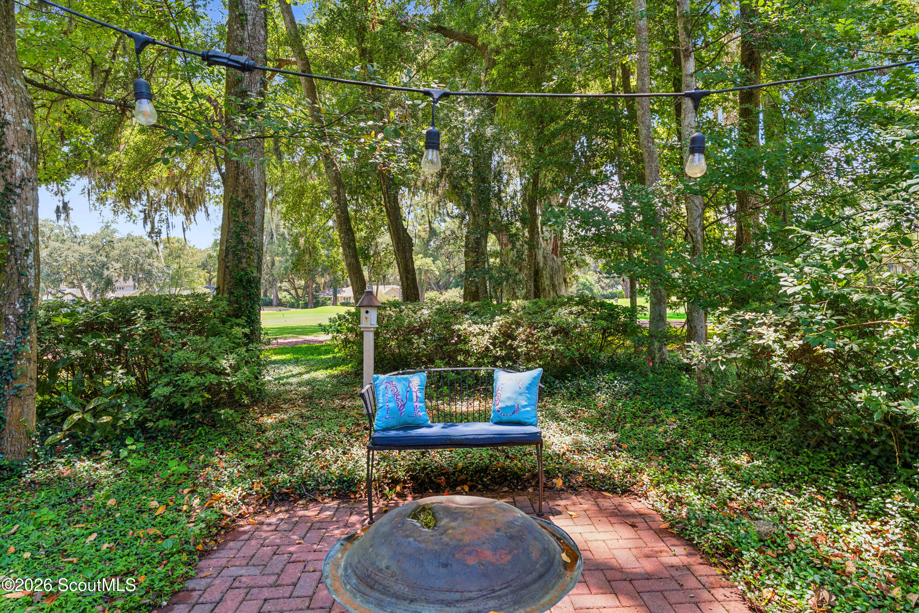 Garden patio with a blue-cushioned bench, metal fire pit, and string lights under mature trees.