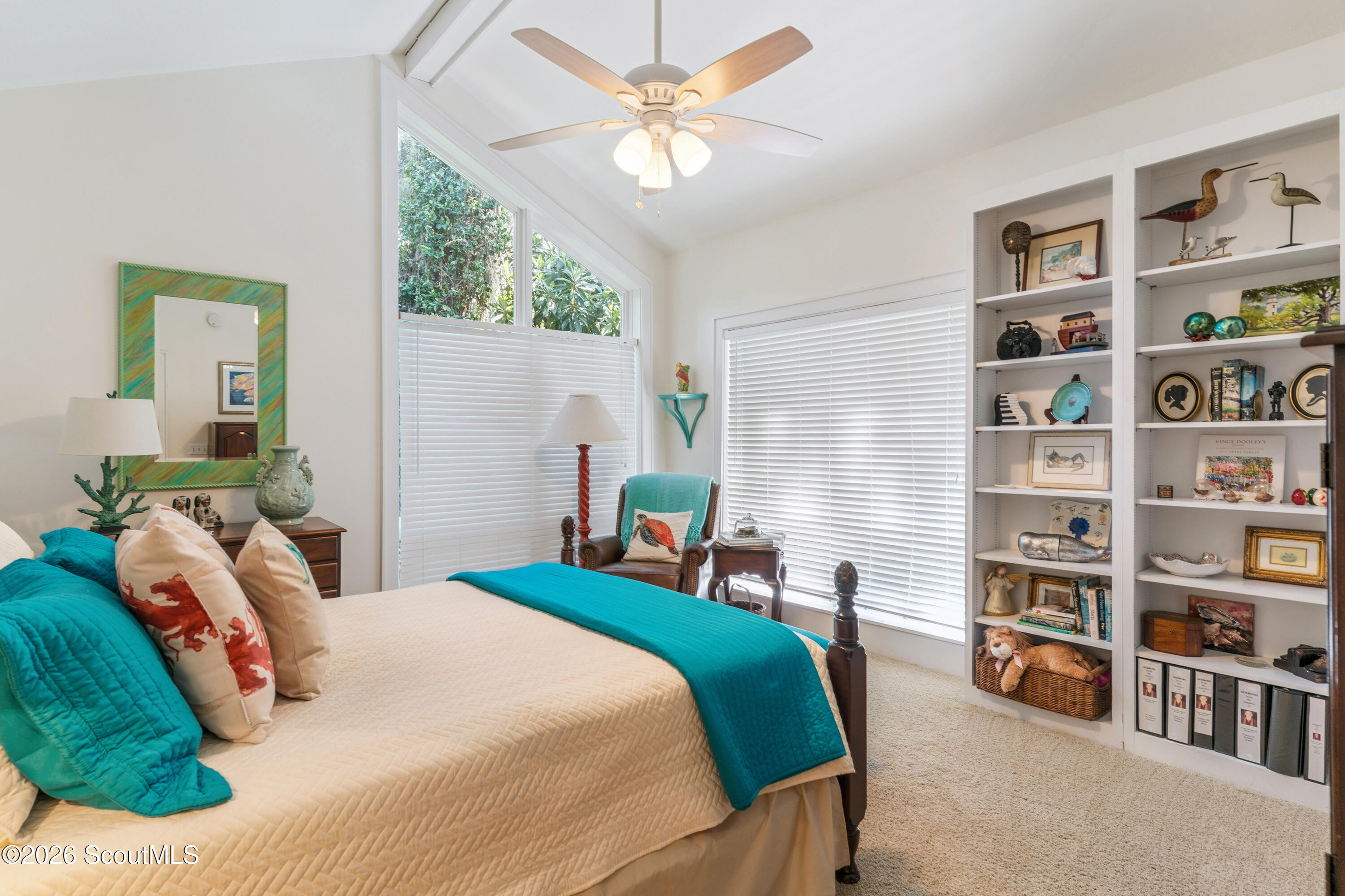 Furnished bedroom with turquoise bedding, tall windows, a reading chair, and built-in display shelves.
