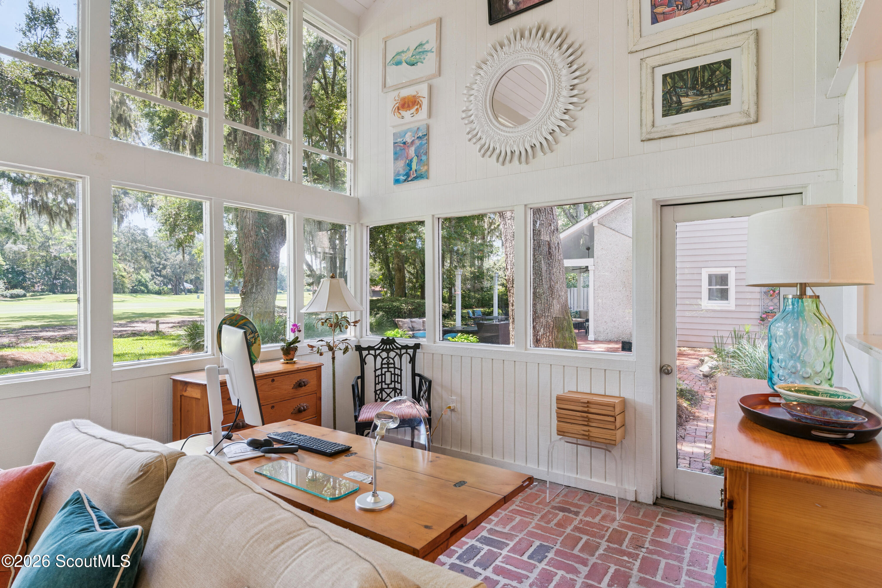 Bright sunroom with tall windows overlooking a lawn, furnished with a sofa and wooden desk.