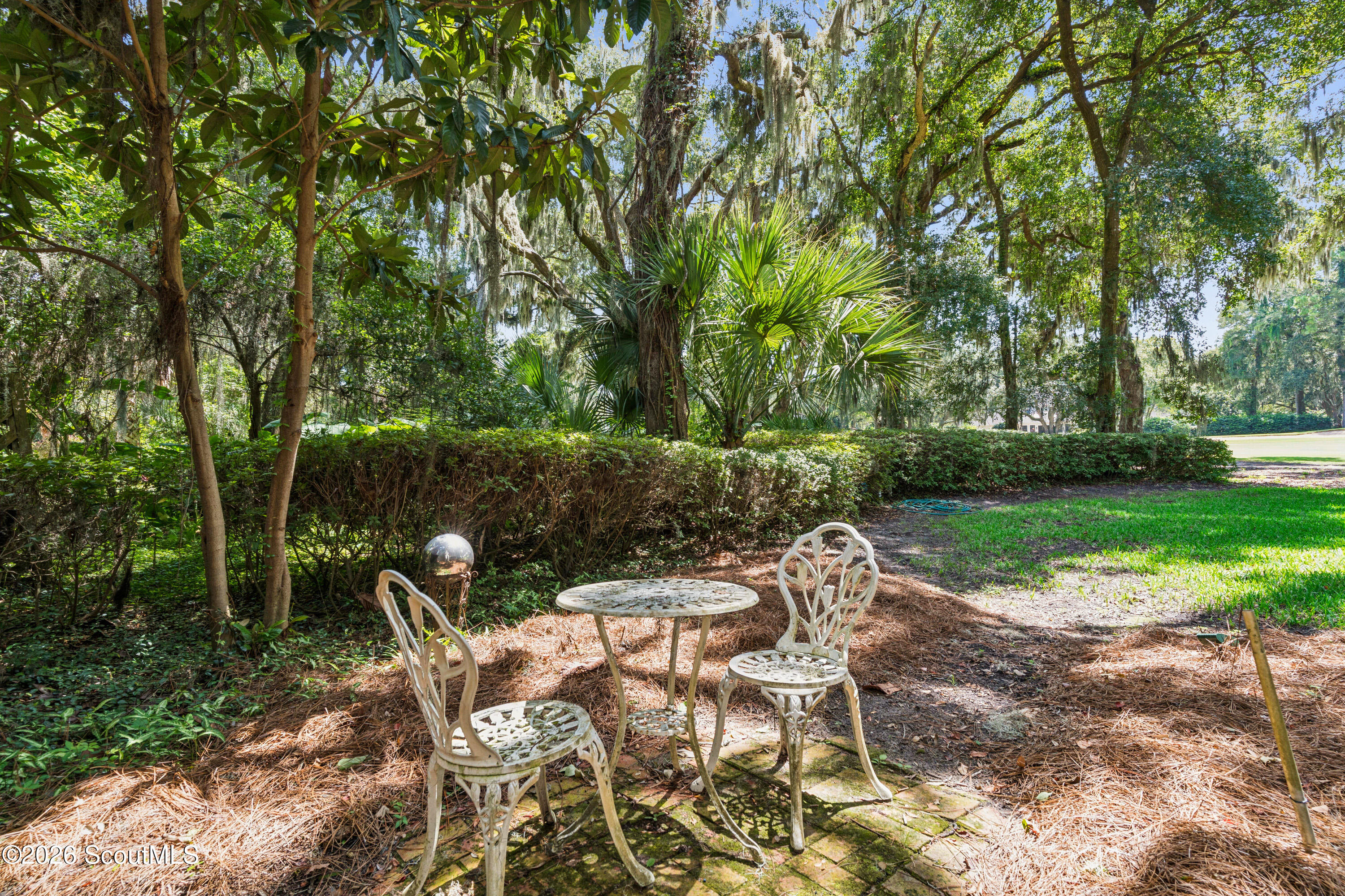 Shaded garden patio with a stone table and white metal chairs beneath palms and moss-draped trees.