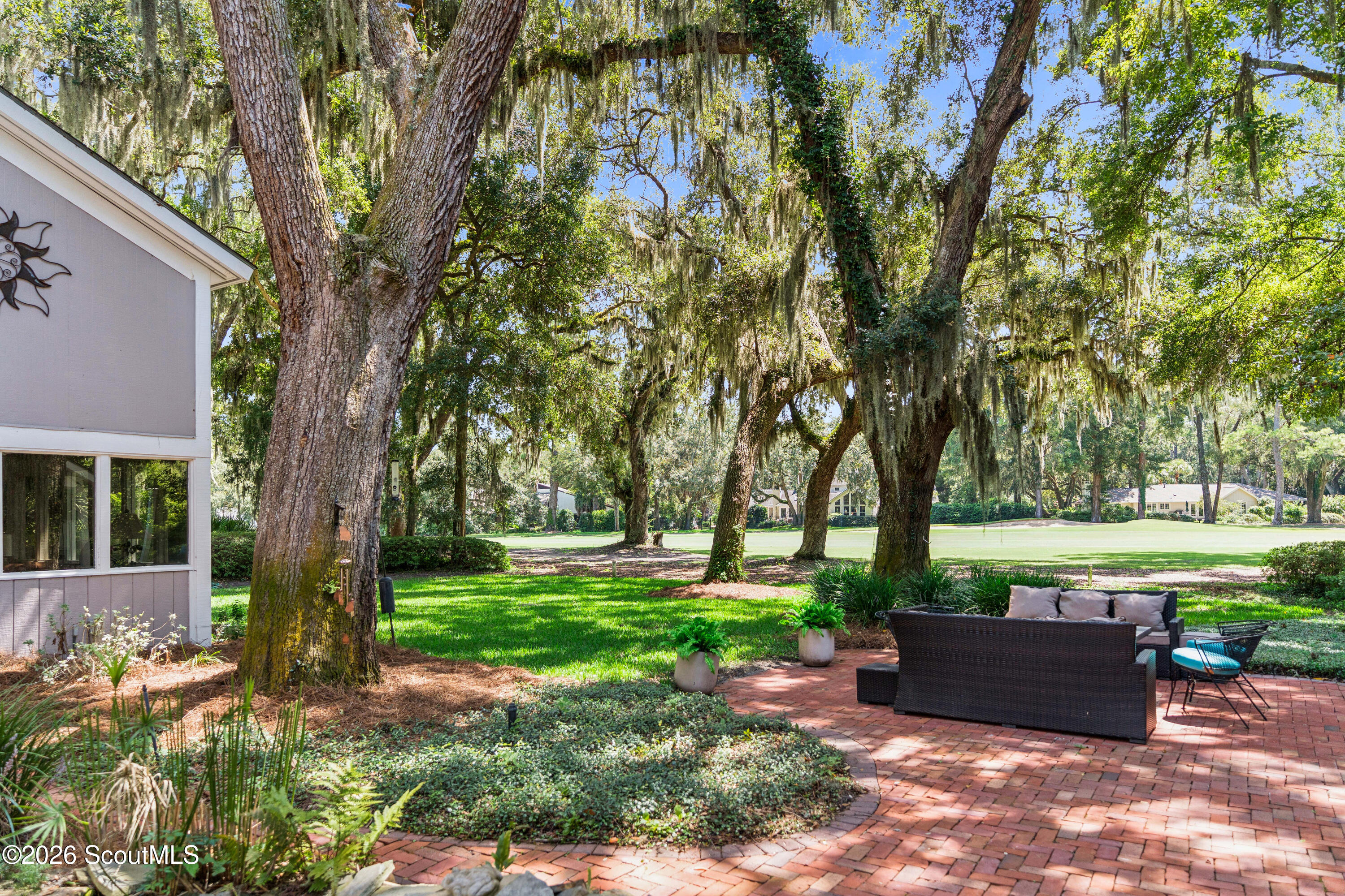 Brick patio with wicker seating beneath moss-draped oak trees beside a broad green lawn.