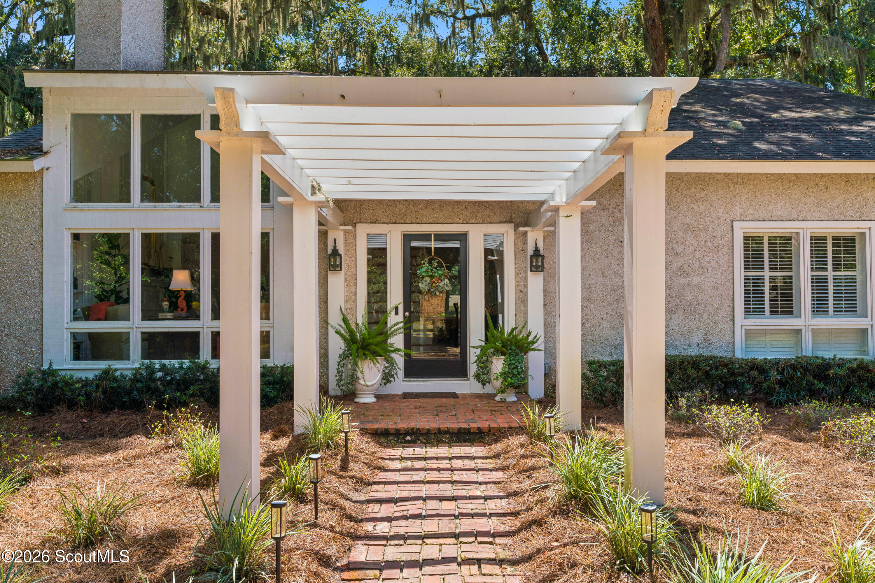 Home entrance beneath a white pergola, with a brick walkway and potted ferns.
