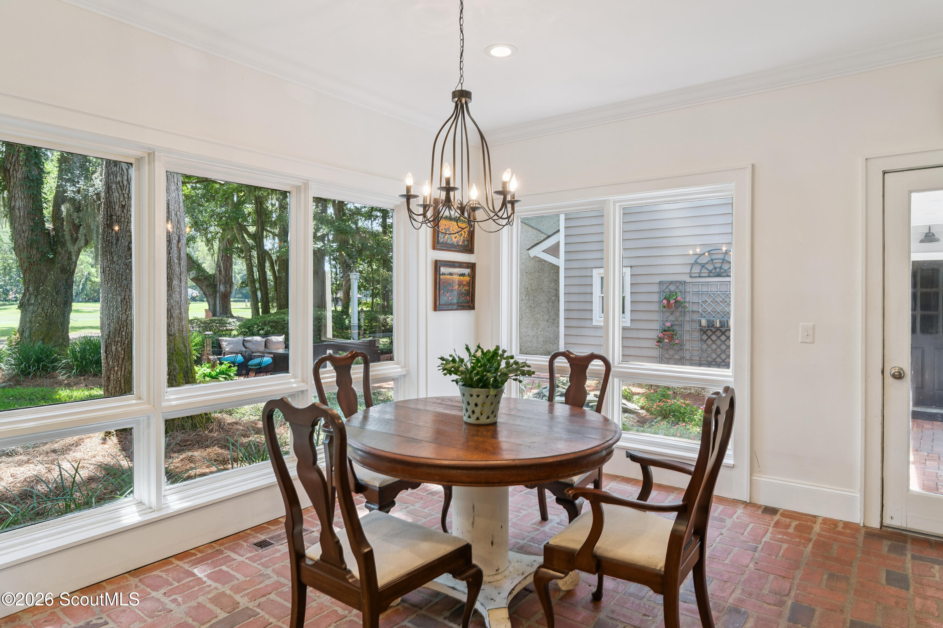Dining room with a round wooden table, brick floor, chandelier, and large garden-facing windows.