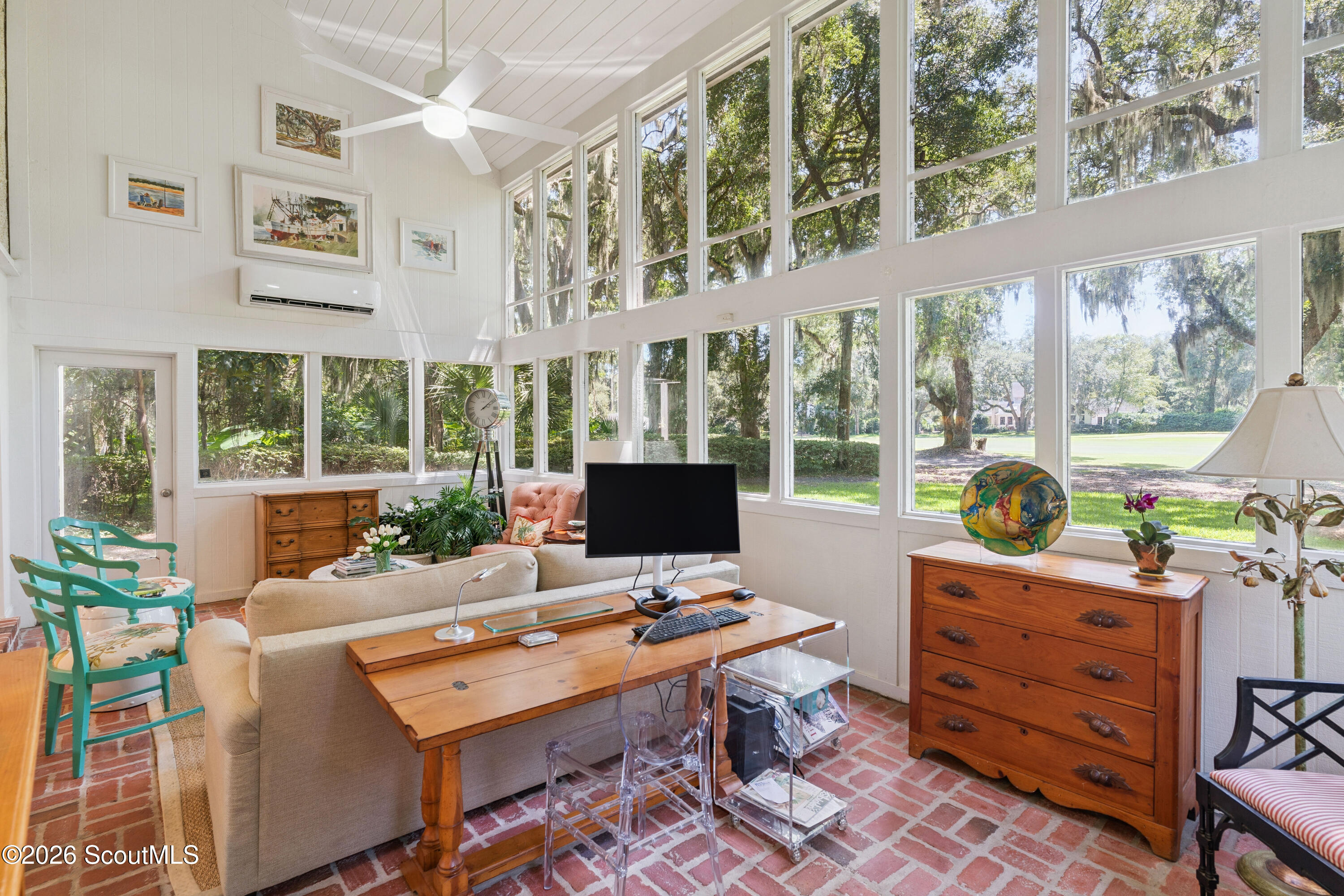 Bright furnished sunroom with expansive windows overlooking a leafy lawn and a red brick floor.