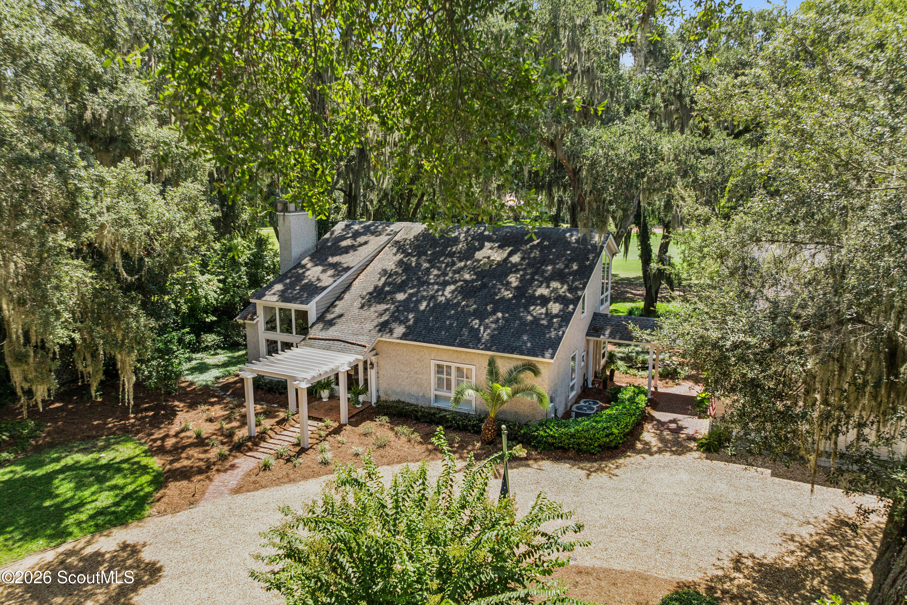 Aerial view of a two-story home framed by mature trees, with a white pergola and gravel drive.