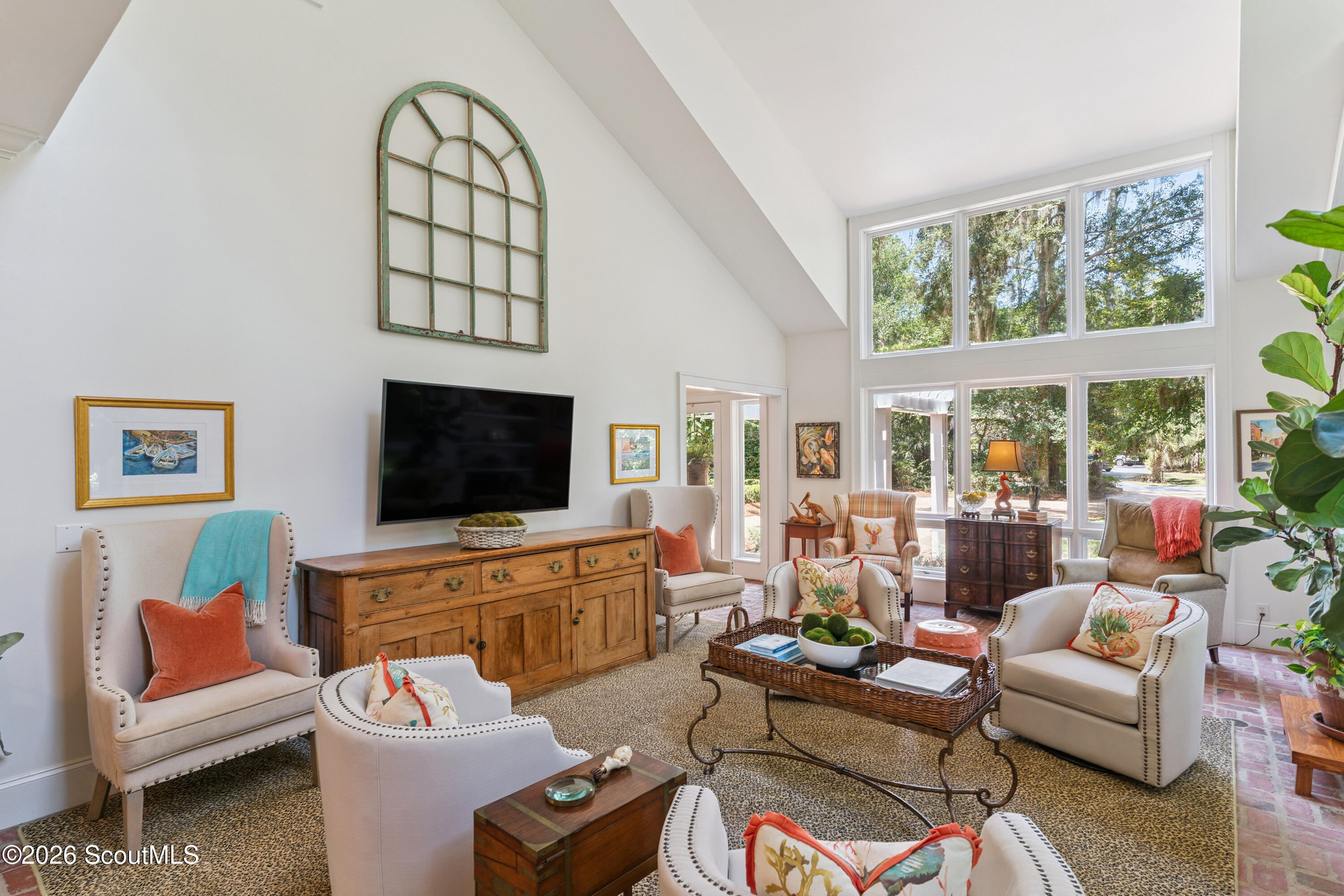 Furnished living room with vaulted ceiling, large leafy-view windows, and cream armchairs around a woven coffee table.