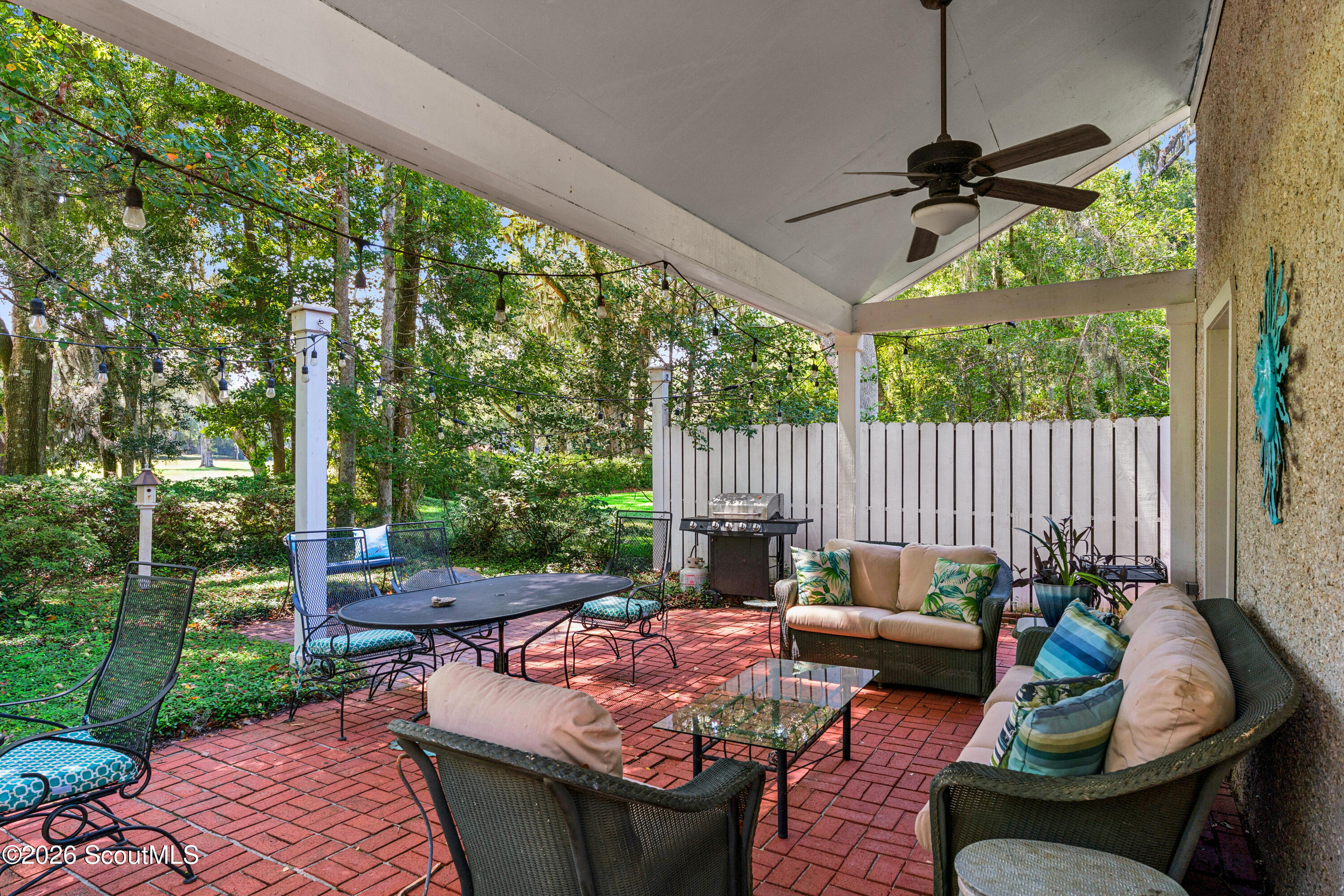 Covered brick patio with wicker seating, string lights, a grill, and a leafy wooded yard.
