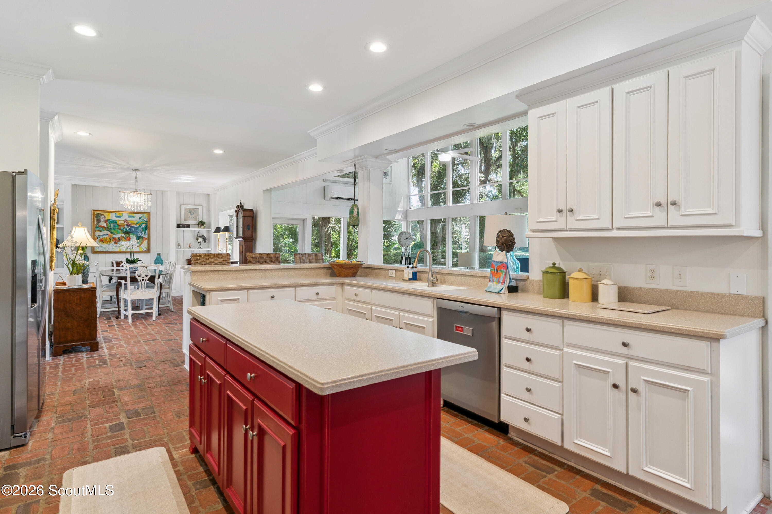 Bright kitchen with a deep red island, brick floors, and large windows overlooking trees.