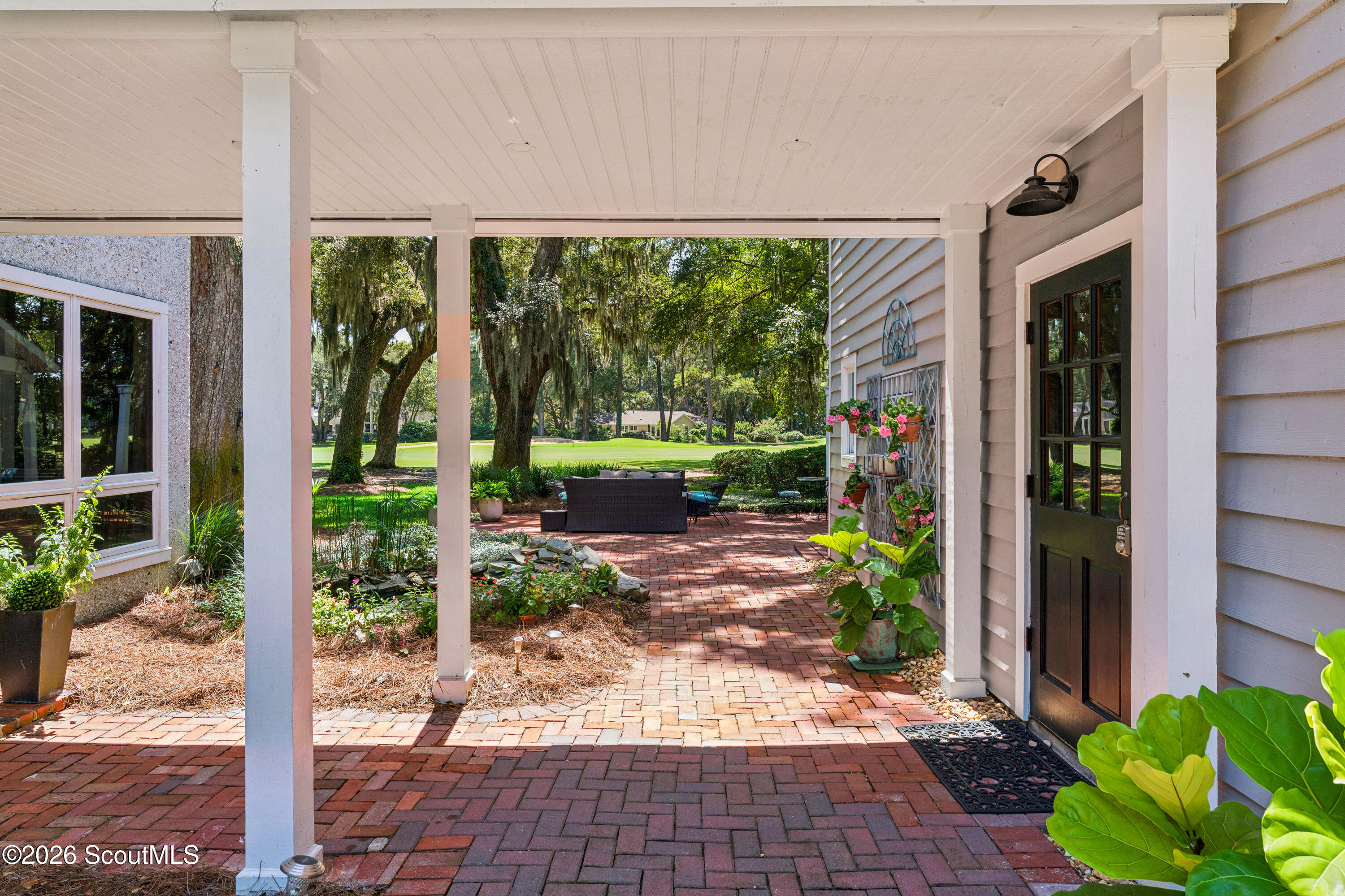 Covered brick patio with white columns, potted flowers, and a seating area beneath mature trees.